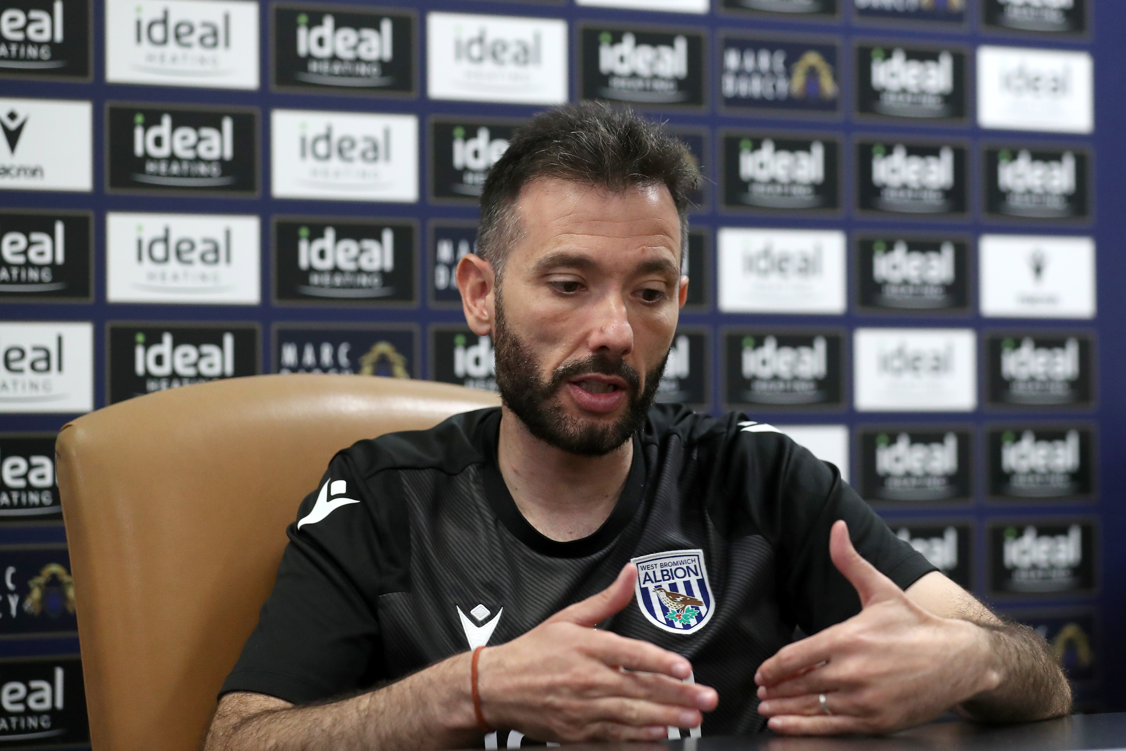 Carlos Corberán speaking at a press conference in front of sponsor boards