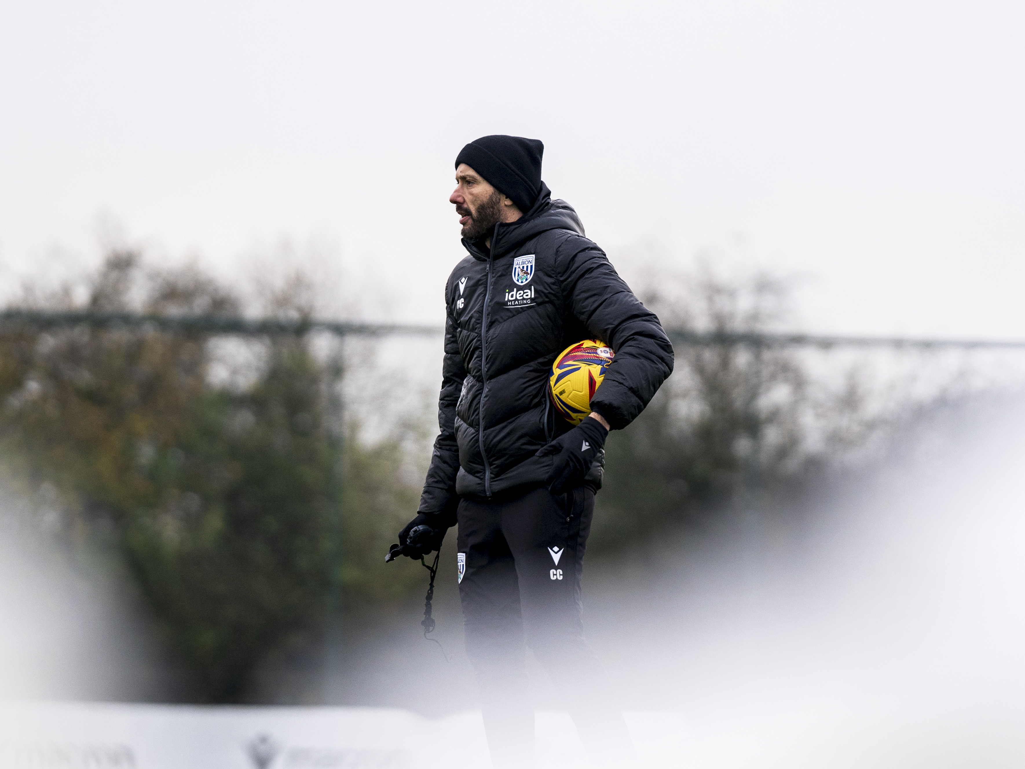 Carlos Corberán watching training while holding a ball and wearing a hat 