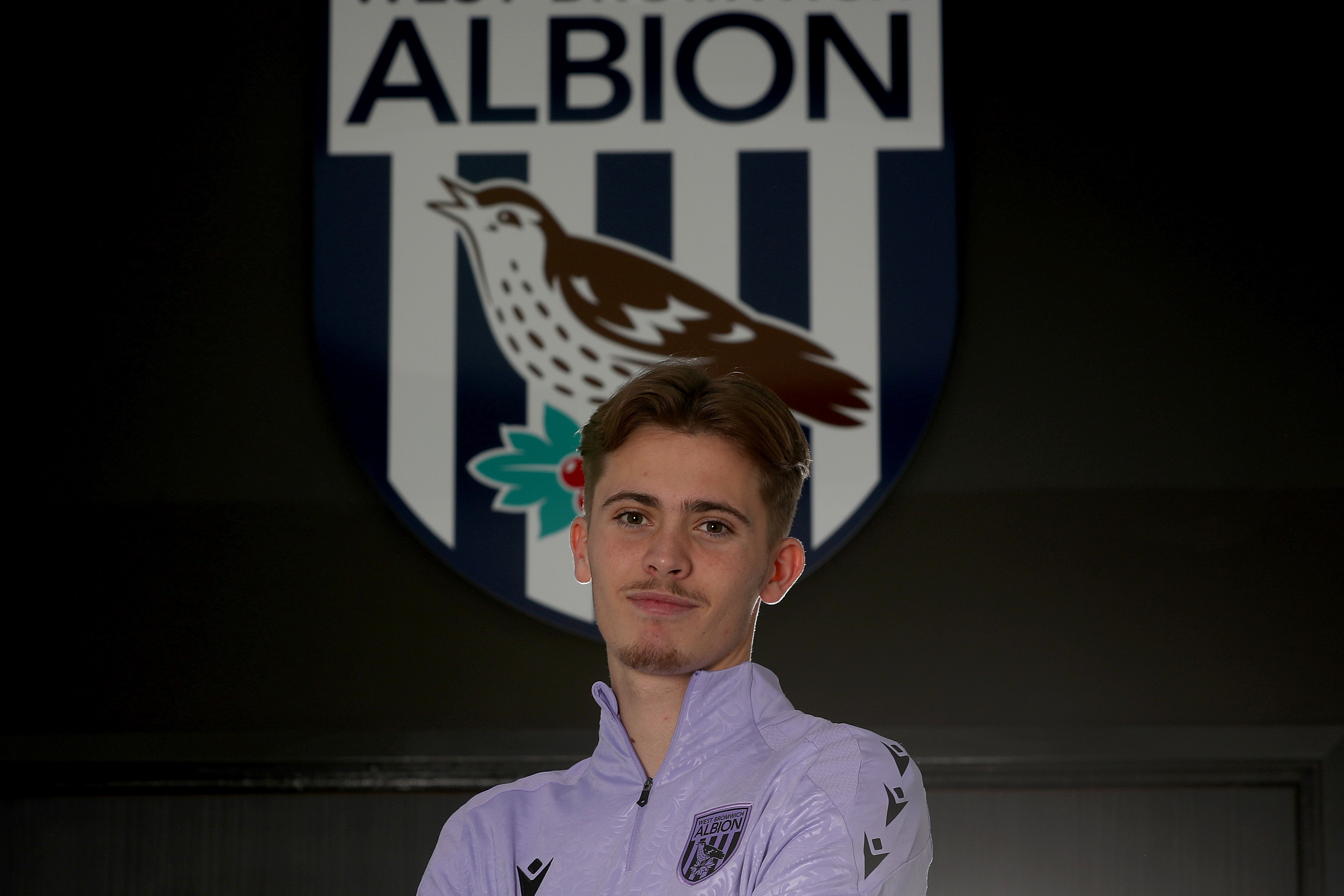 Isaac Price looking at the camera stood in front of a WBA badge 