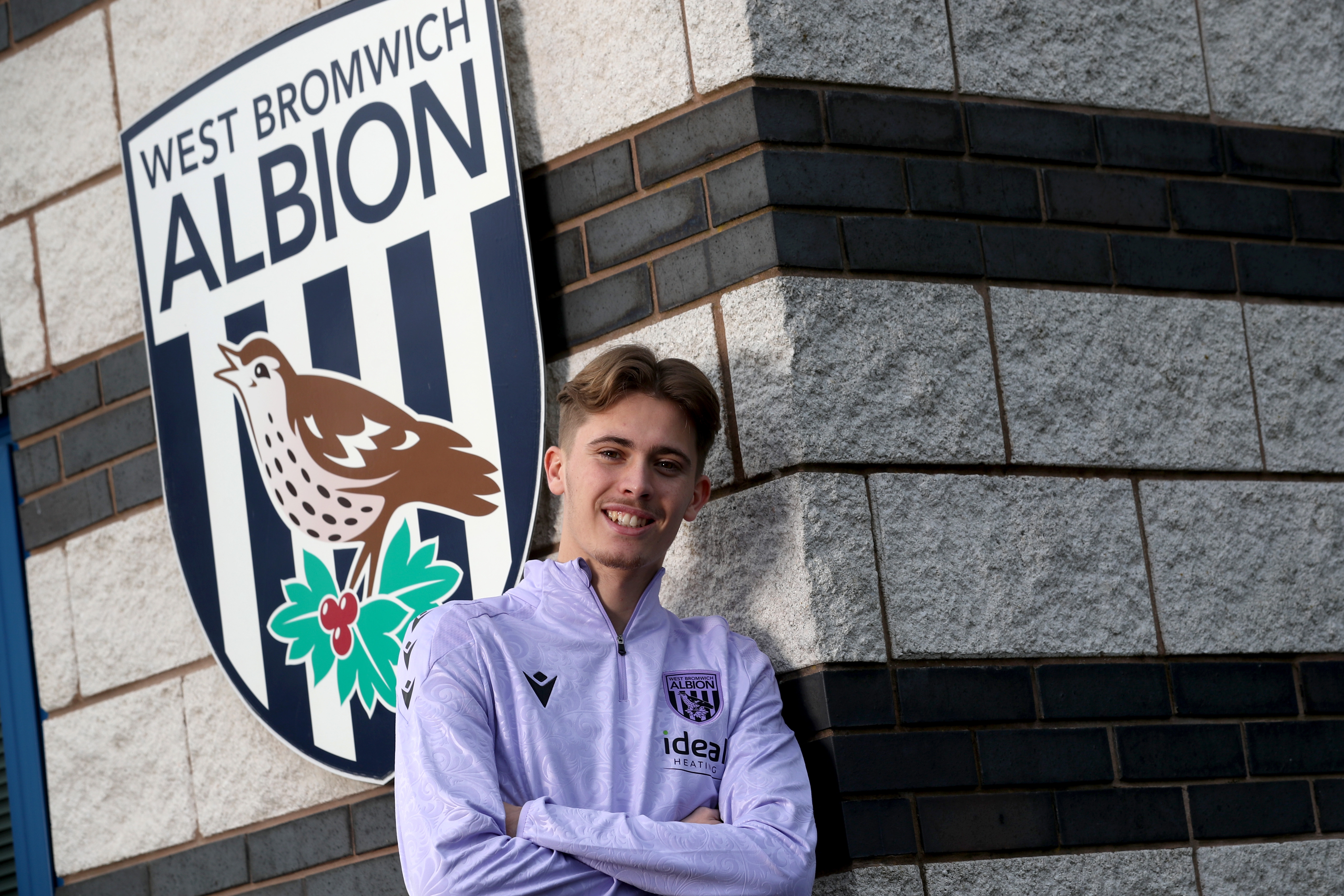 Isaac Price stood up against a wall by a big WBA badge with his arms crossed 