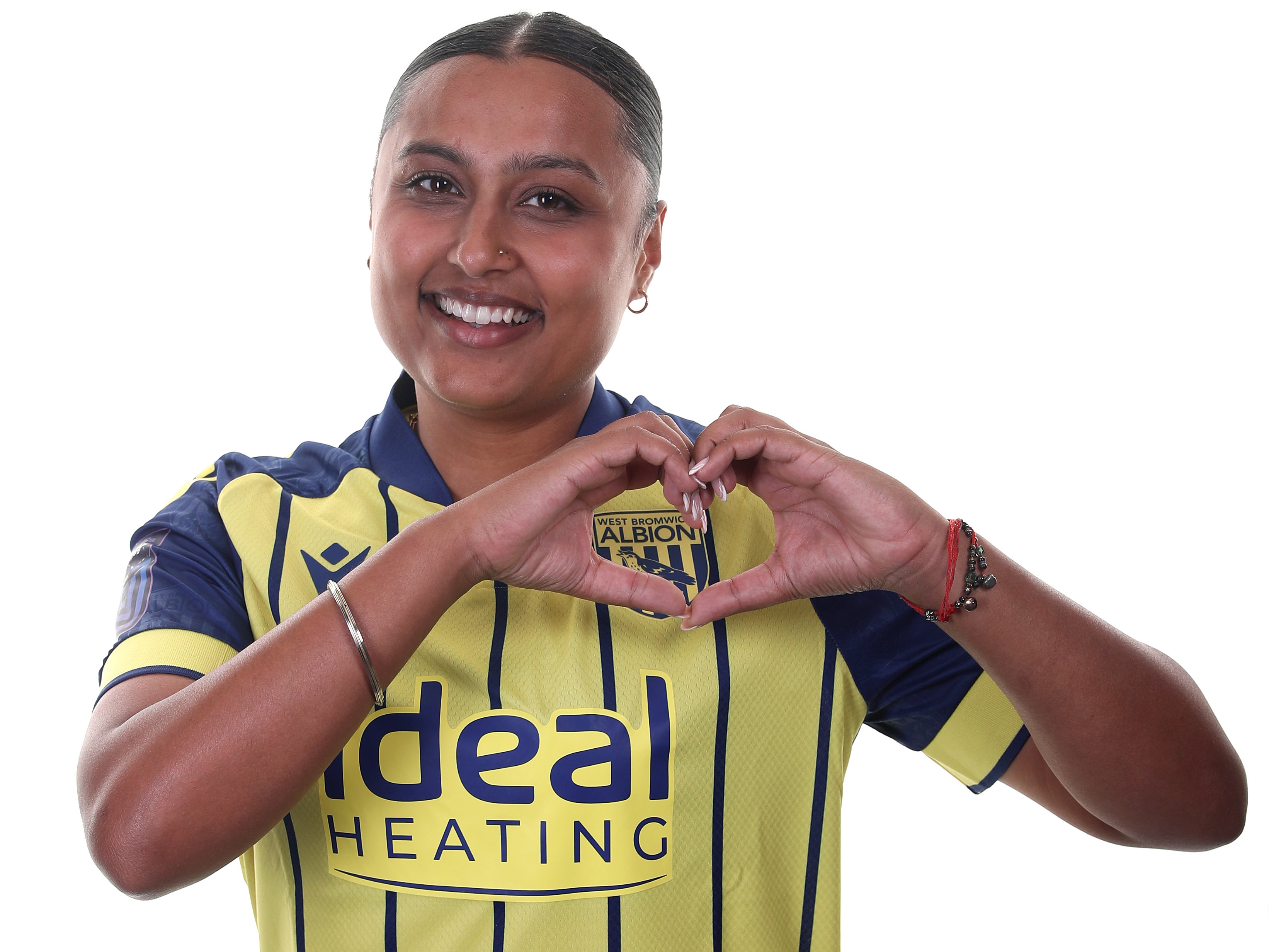 Simran Jhamat smiling at the camera and making a heart shape with her hands while stood in the yellow and navy blue away kit 