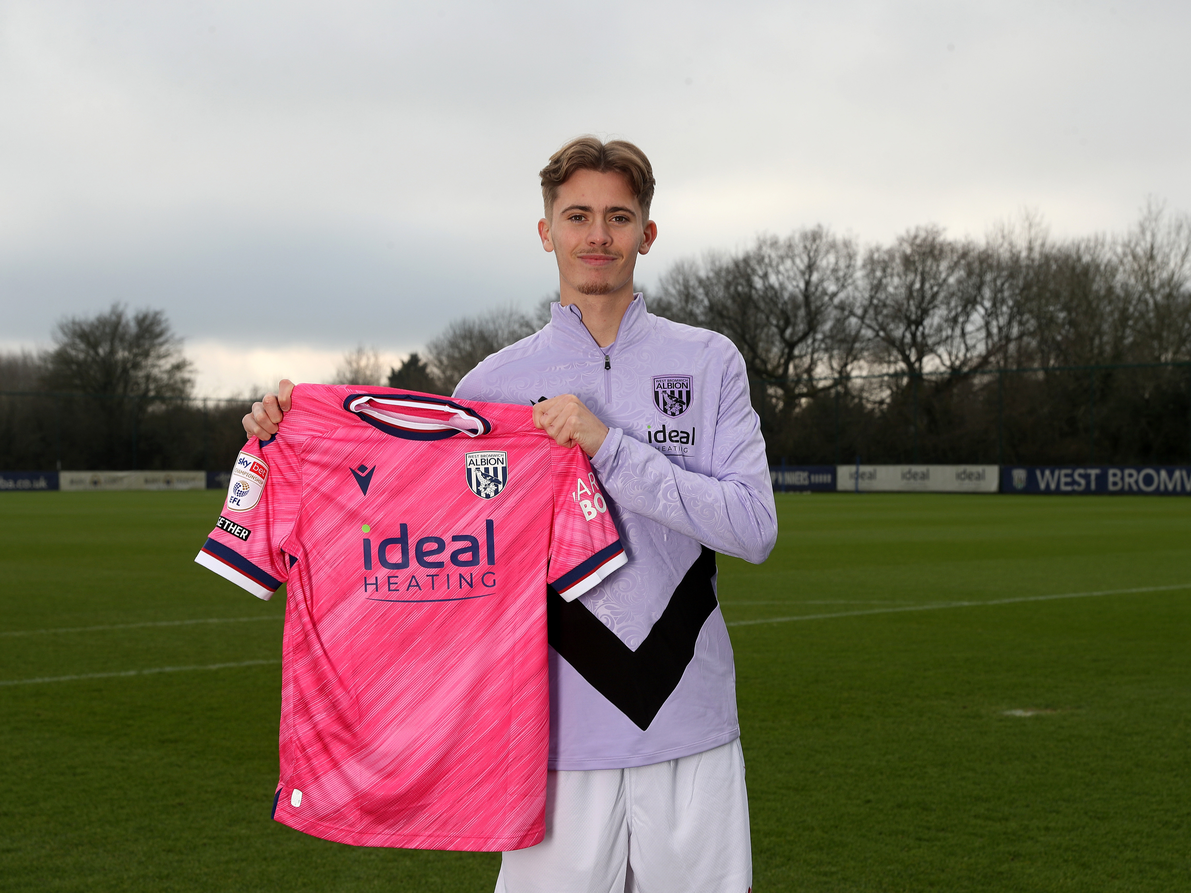 Isaac Price holding up a pink WBA shirt and smiling at the camera 