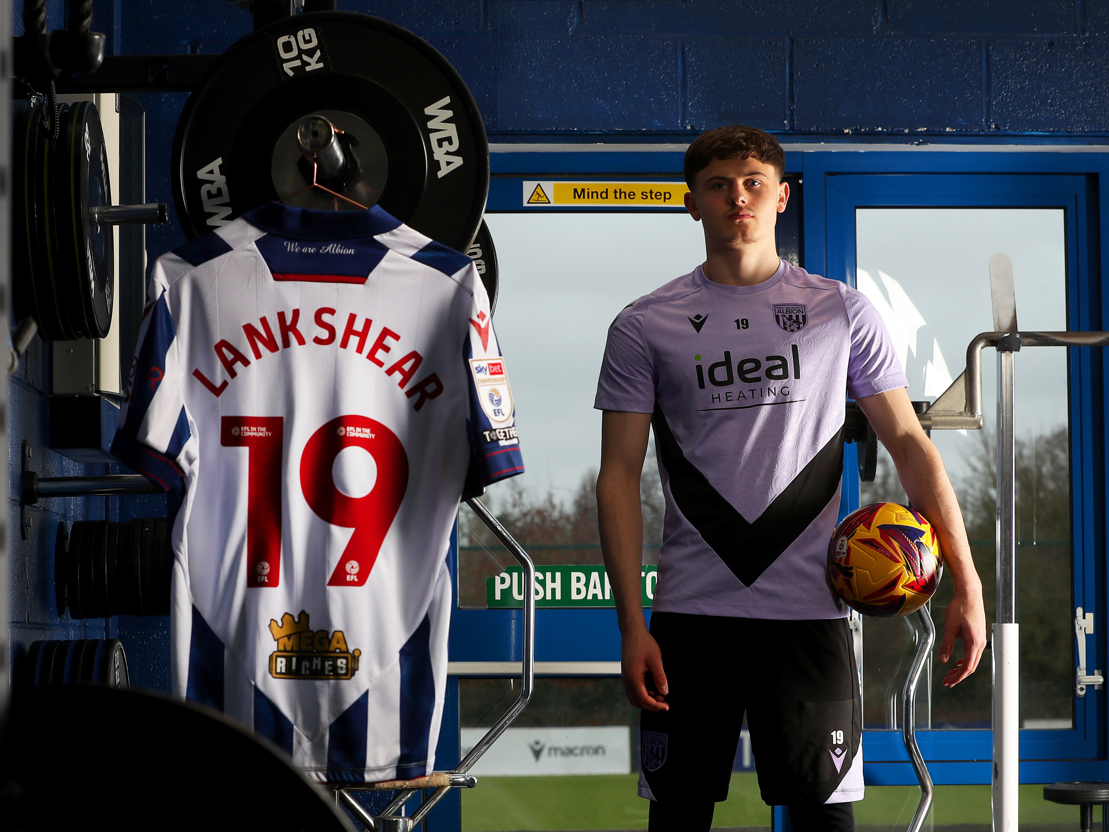 Will Lankshear standing in the gym holding a ball next to his shirt hanging up with his name and number 19 on the back