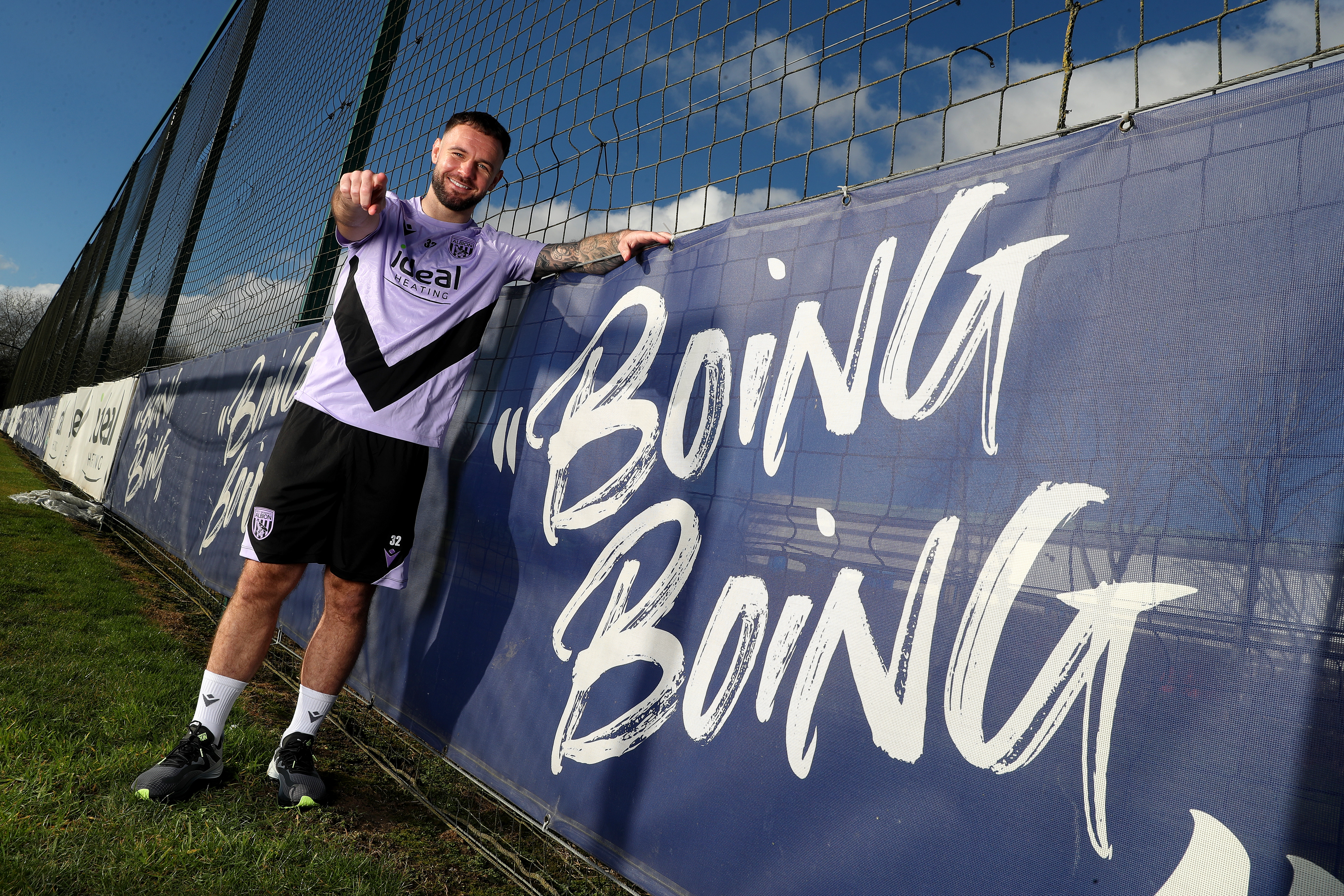 Adam Armstrong smiling and pointing at the camera while stood next to branding round the side of a pitch with "boing boing" written on