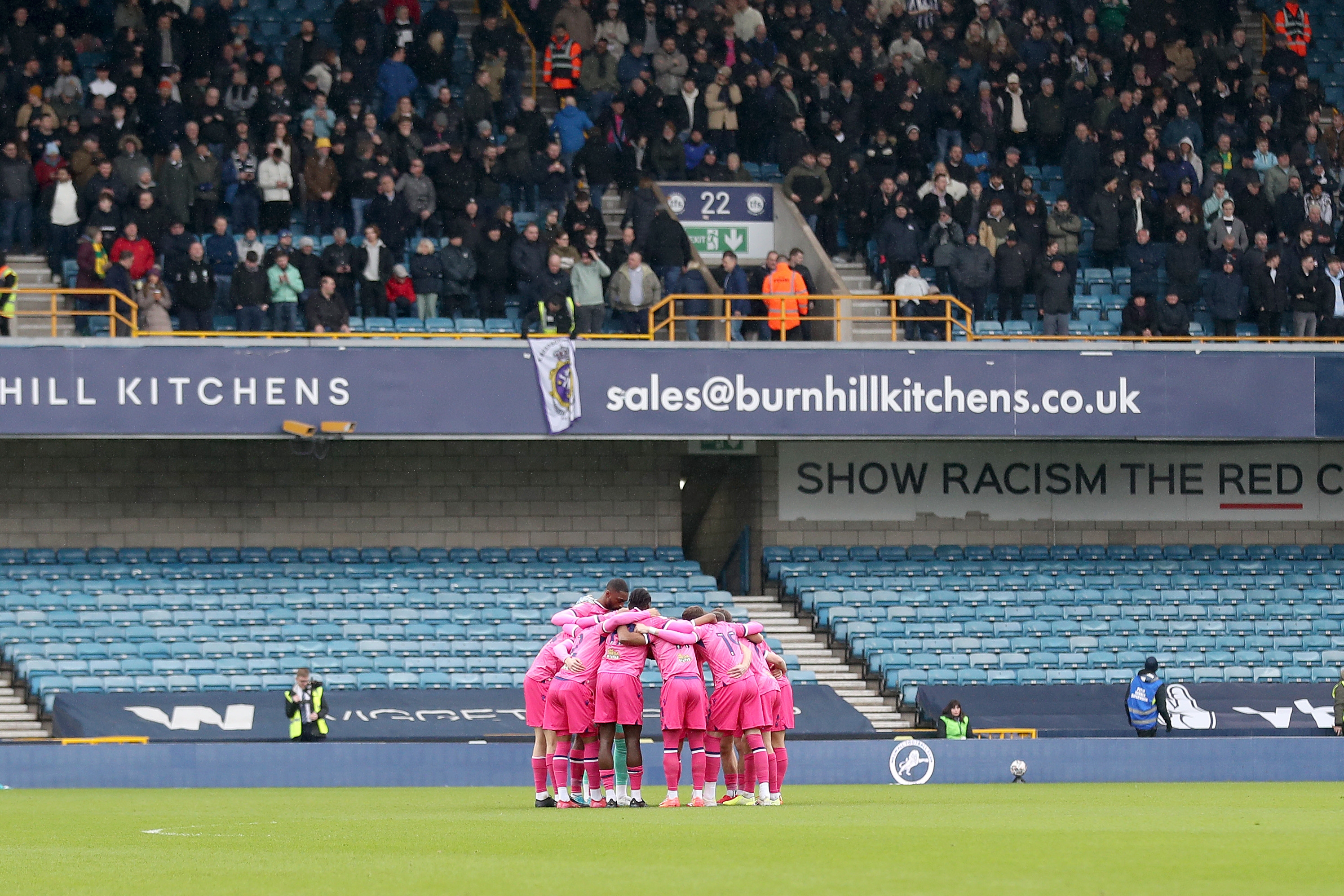 Albion players in the pink kit in a pre-match huddle at Millwall with the away fans in the background 