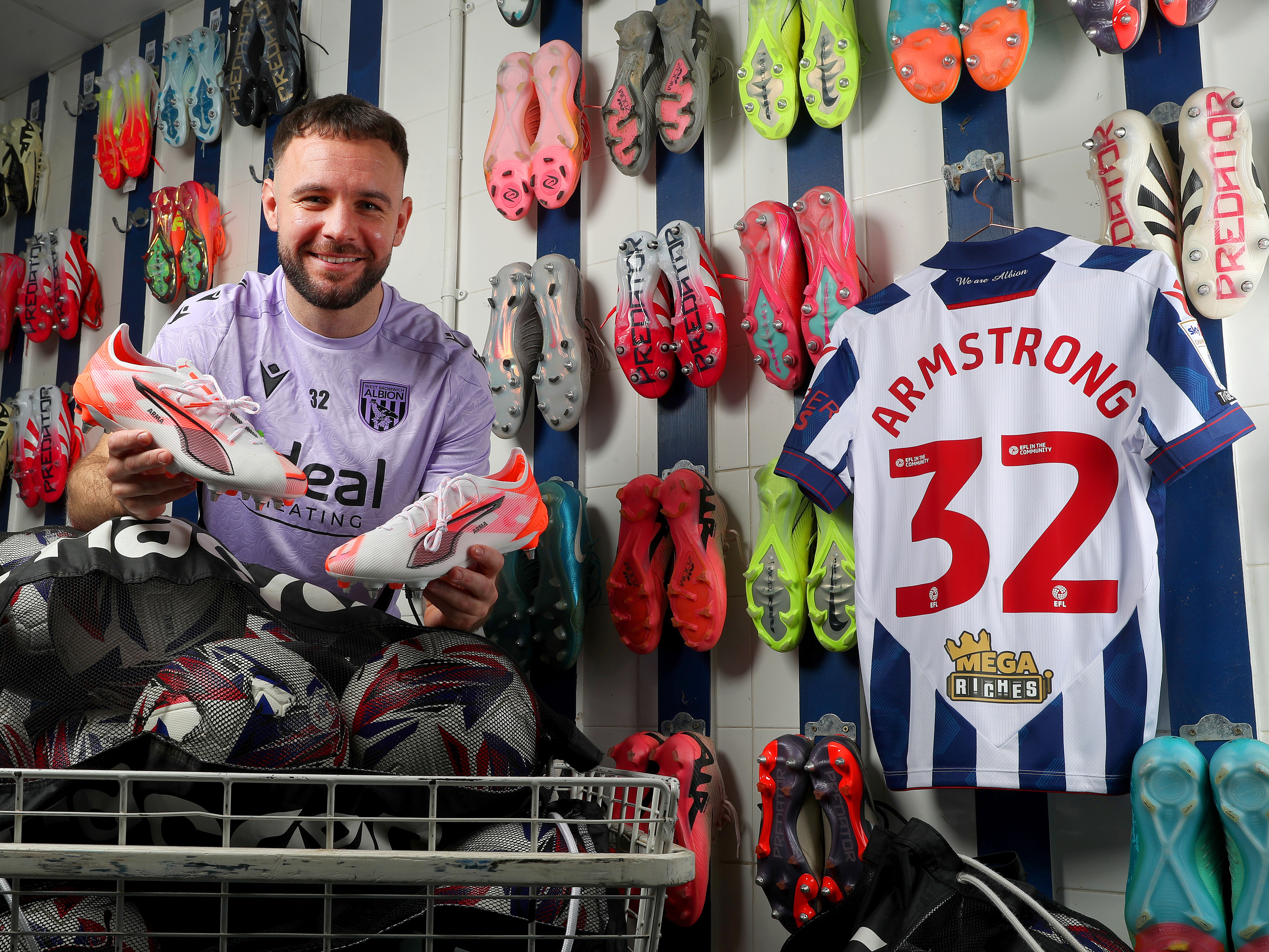 Adam Armstrong smiling at the camera holding his boots while sat in the boot room with his home shirt hanging up back-to-front with his number 32 on show