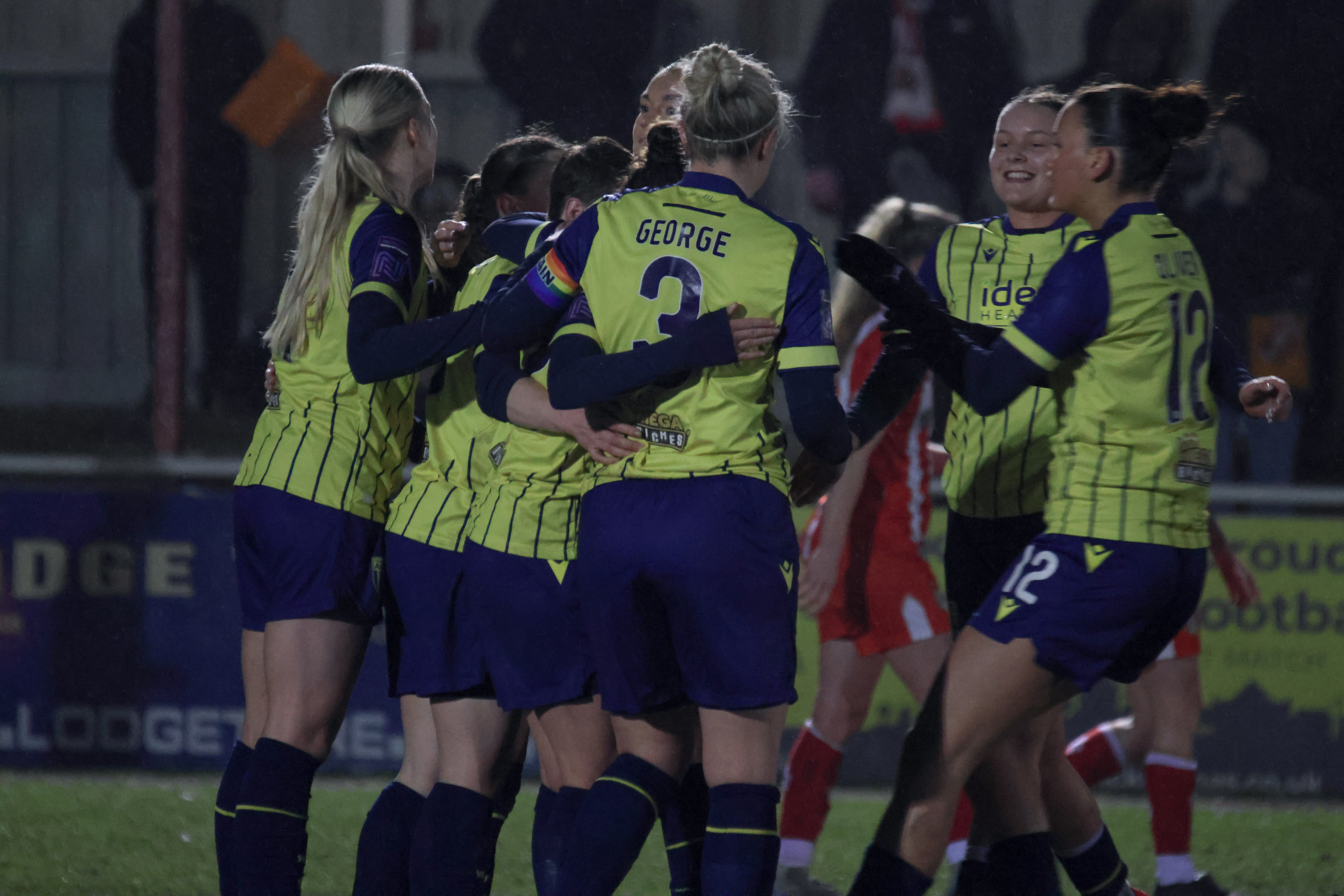 Hannah George in the yellow and navy blue away kit celebrating with team-mates after a goal 