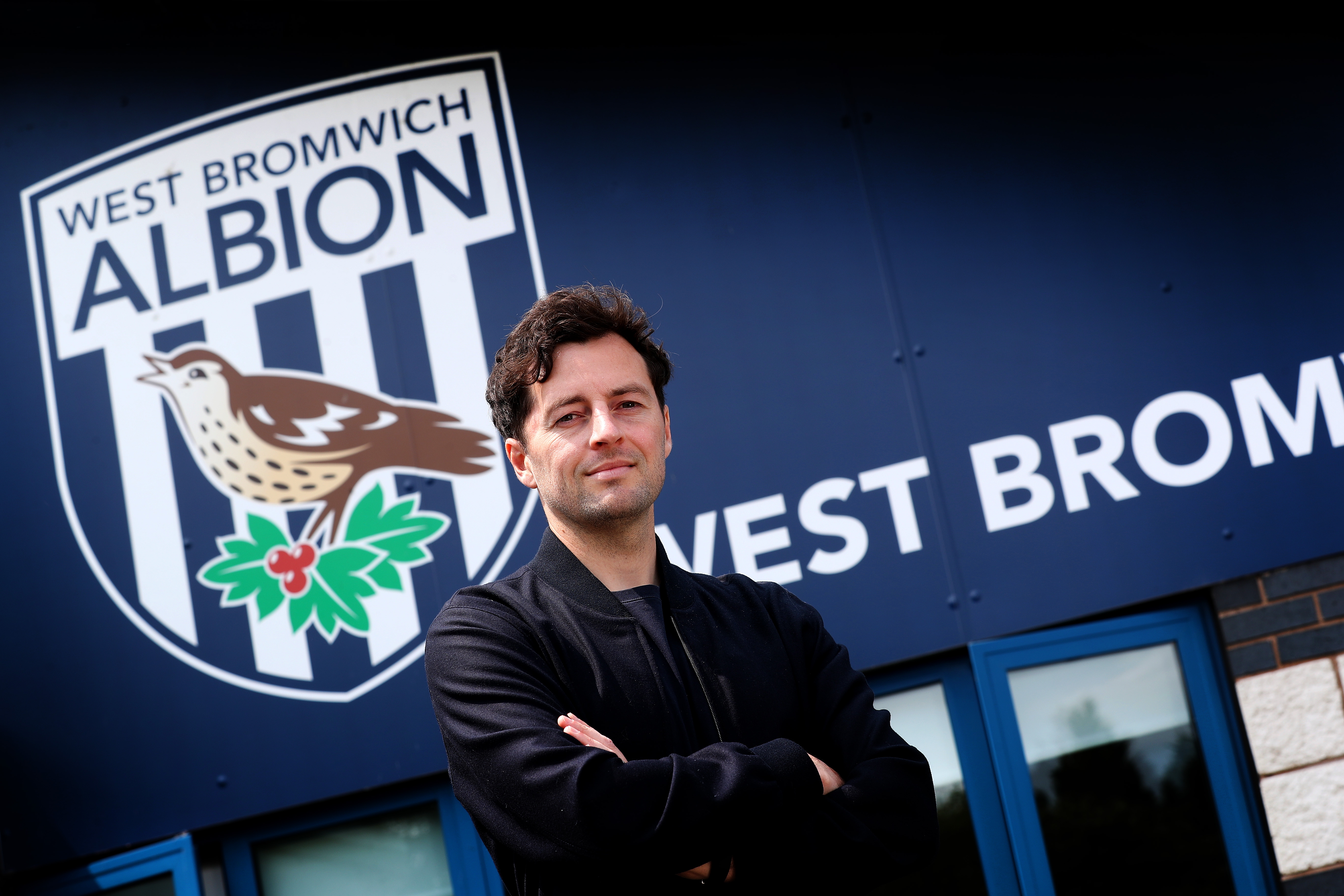 Ryan Mason posing for a photo with his arms crossed and a smile on his face looking at the camera while stood in front of WBA branding
