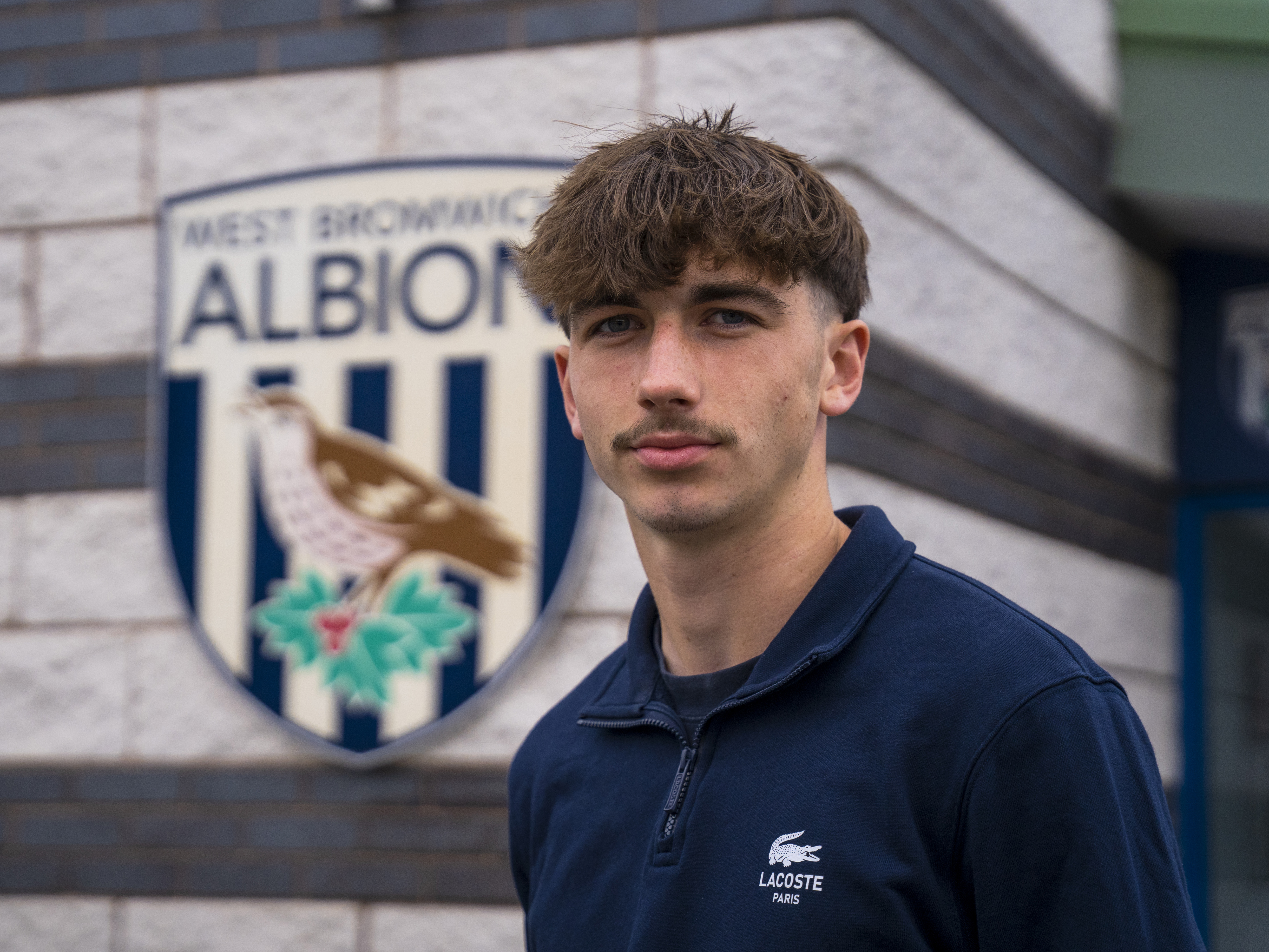 A photo of Albion youngster Alfie Maughan standing in front of the Albion badge at the front of the training ground