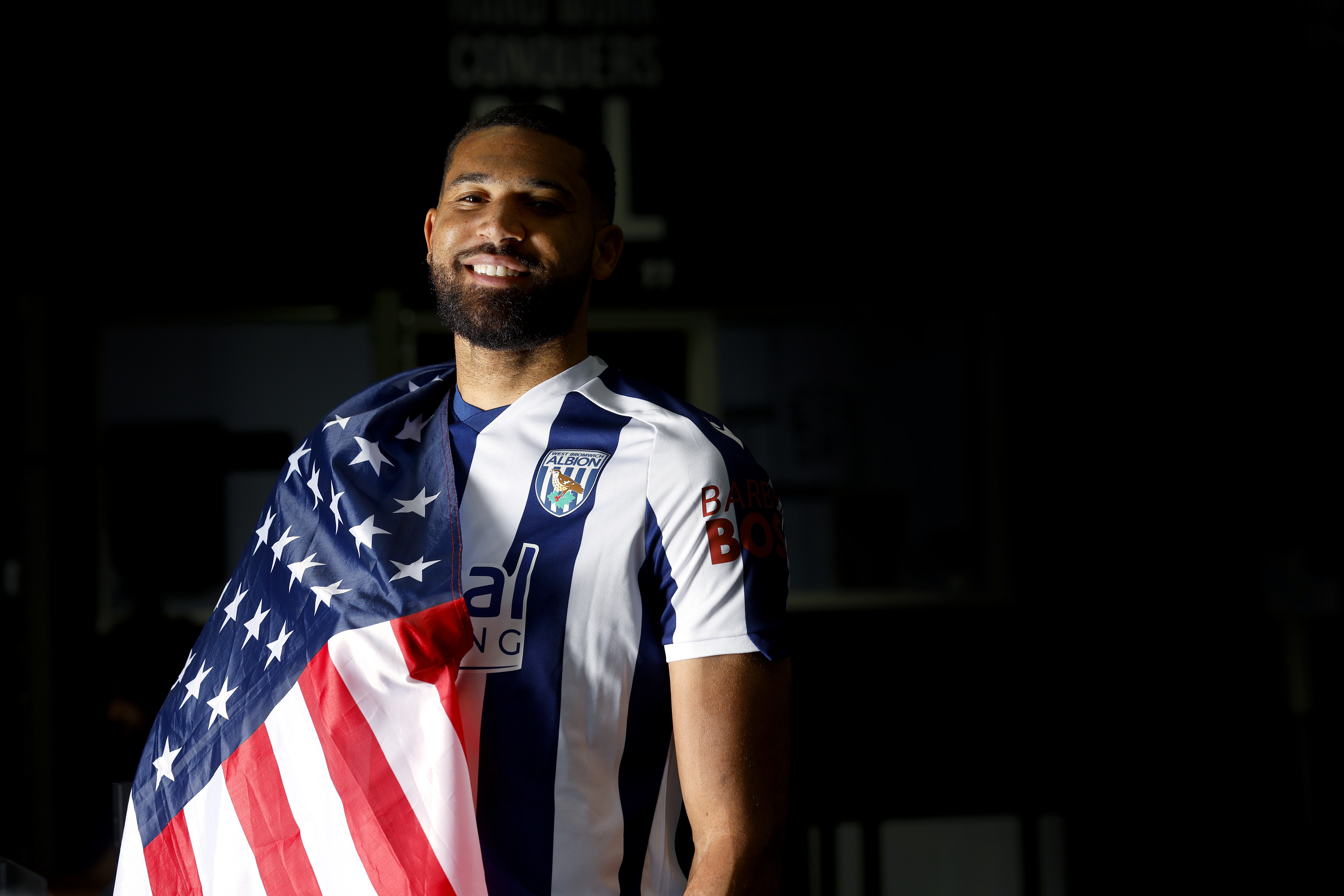 George Campbell smiling at the camera while in a home WBA shirt with a USA flag wrapped around his shoulder
