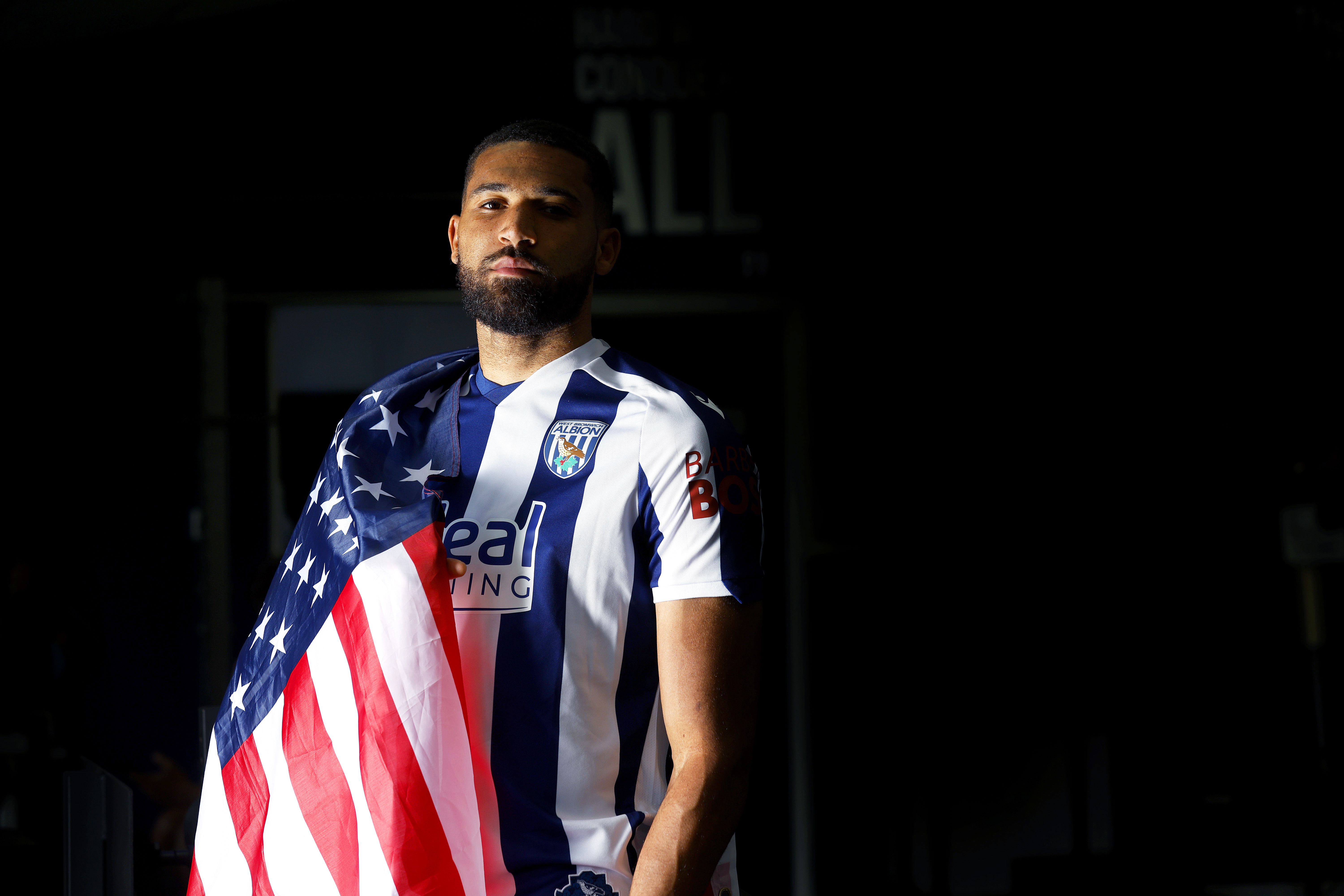 George Campbell looking at the camera while in a home WBA shirt with a USA flag wrapped around his shoulder