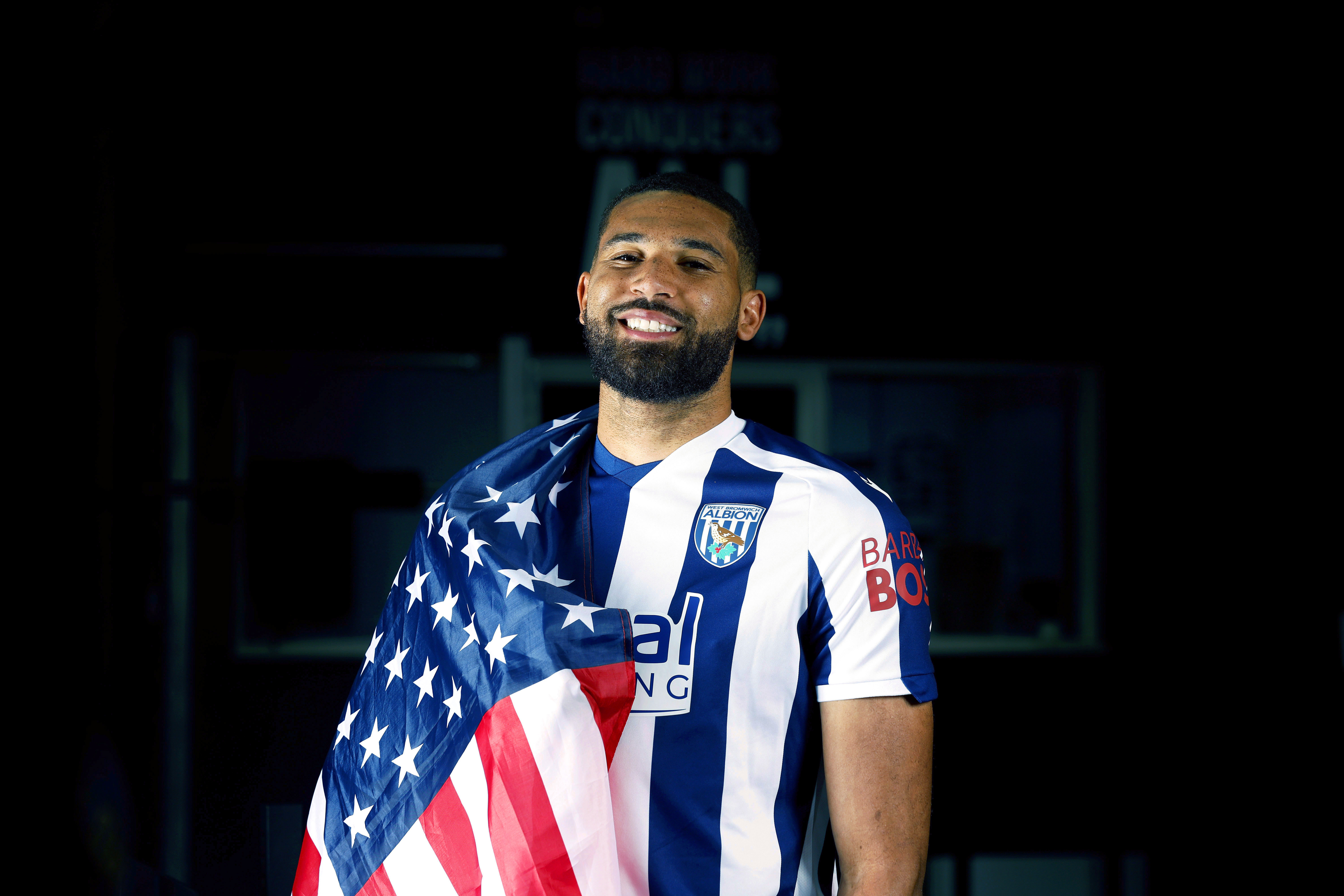 George Campbell smiling at the camera while in a home WBA shirt with a USA flag wrapped around his shoulder