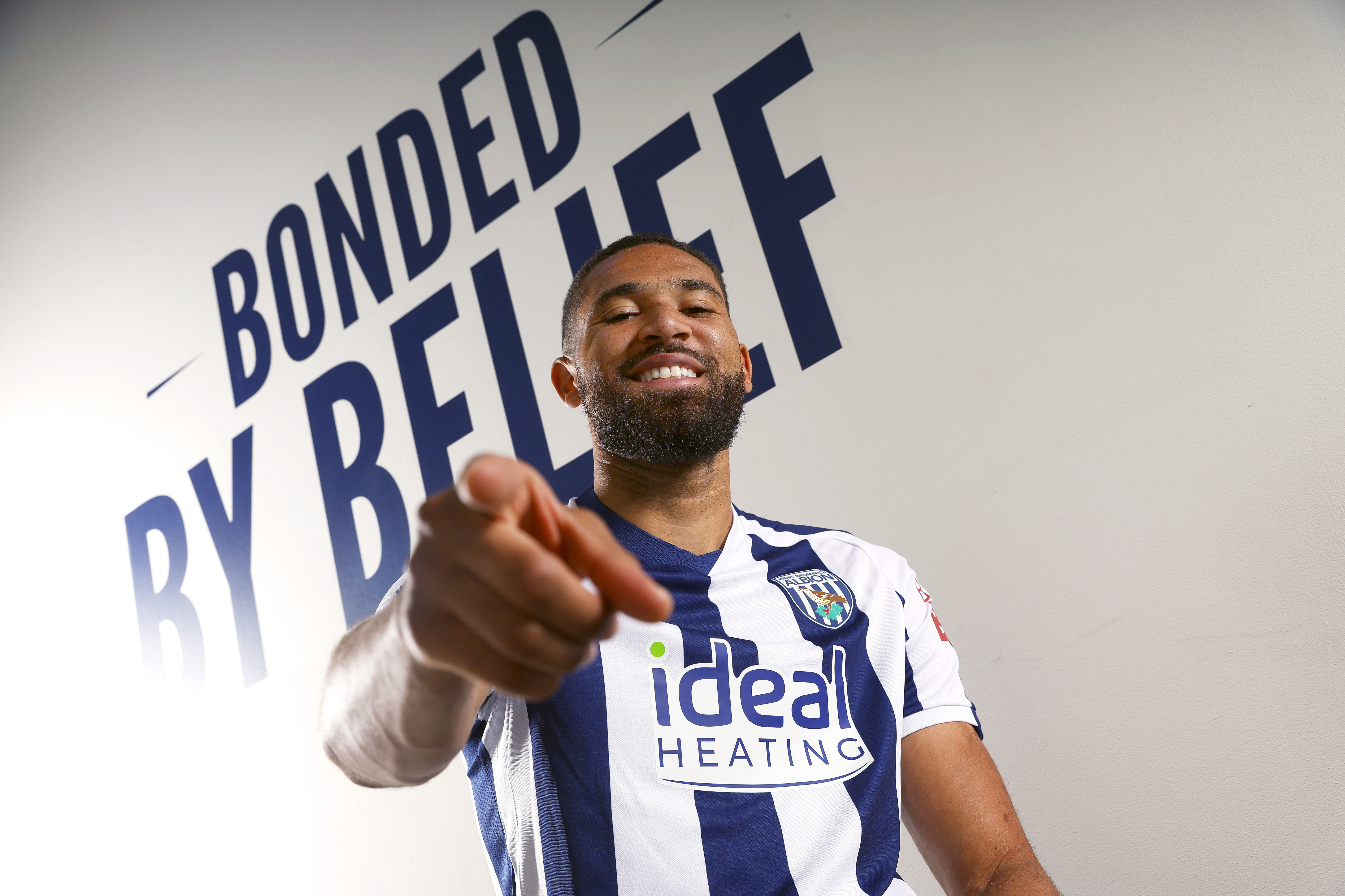 George Campbell smiling and pointing at the camera while sat in a WBA home shirt in front of branding which reads 'bonded by belief'