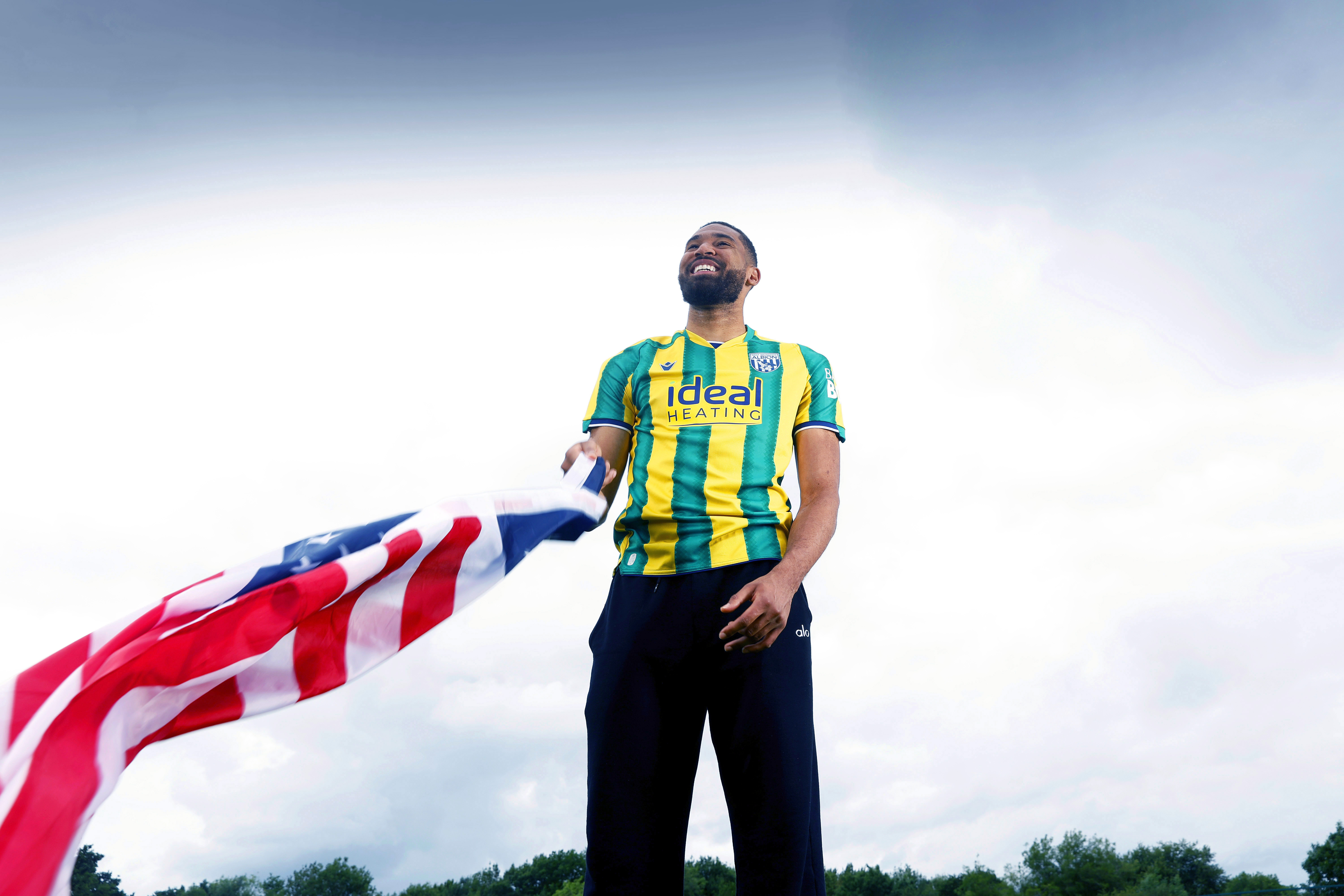 George Campbell smiling while stood in an away WBA shirt waving an American flag