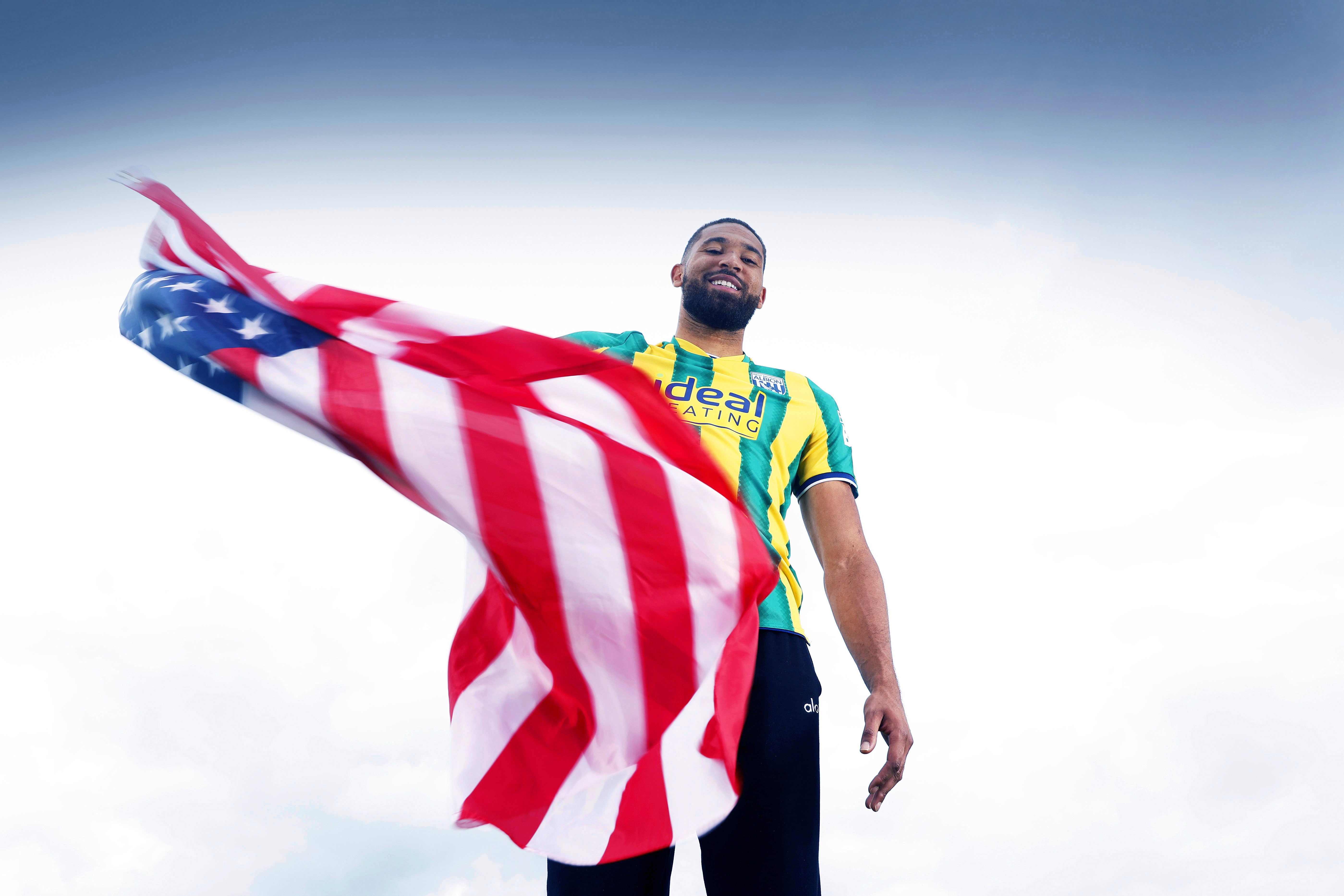 George Campbell smiling while stood in an away WBA shirt waving an American flag
