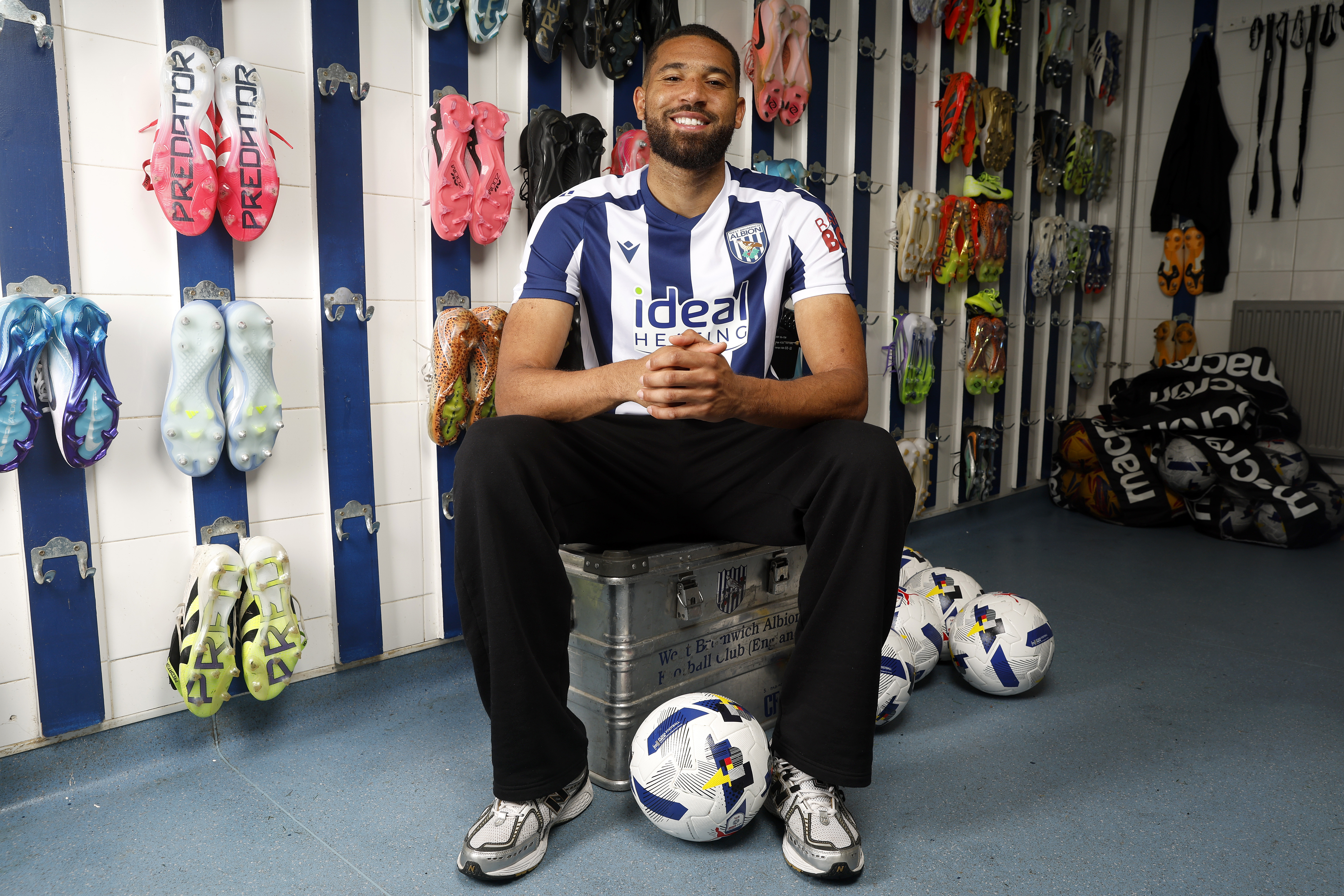 George Campbell sat in the boot room in a home WBA shirt smiling at the camera with footballs by his feet 