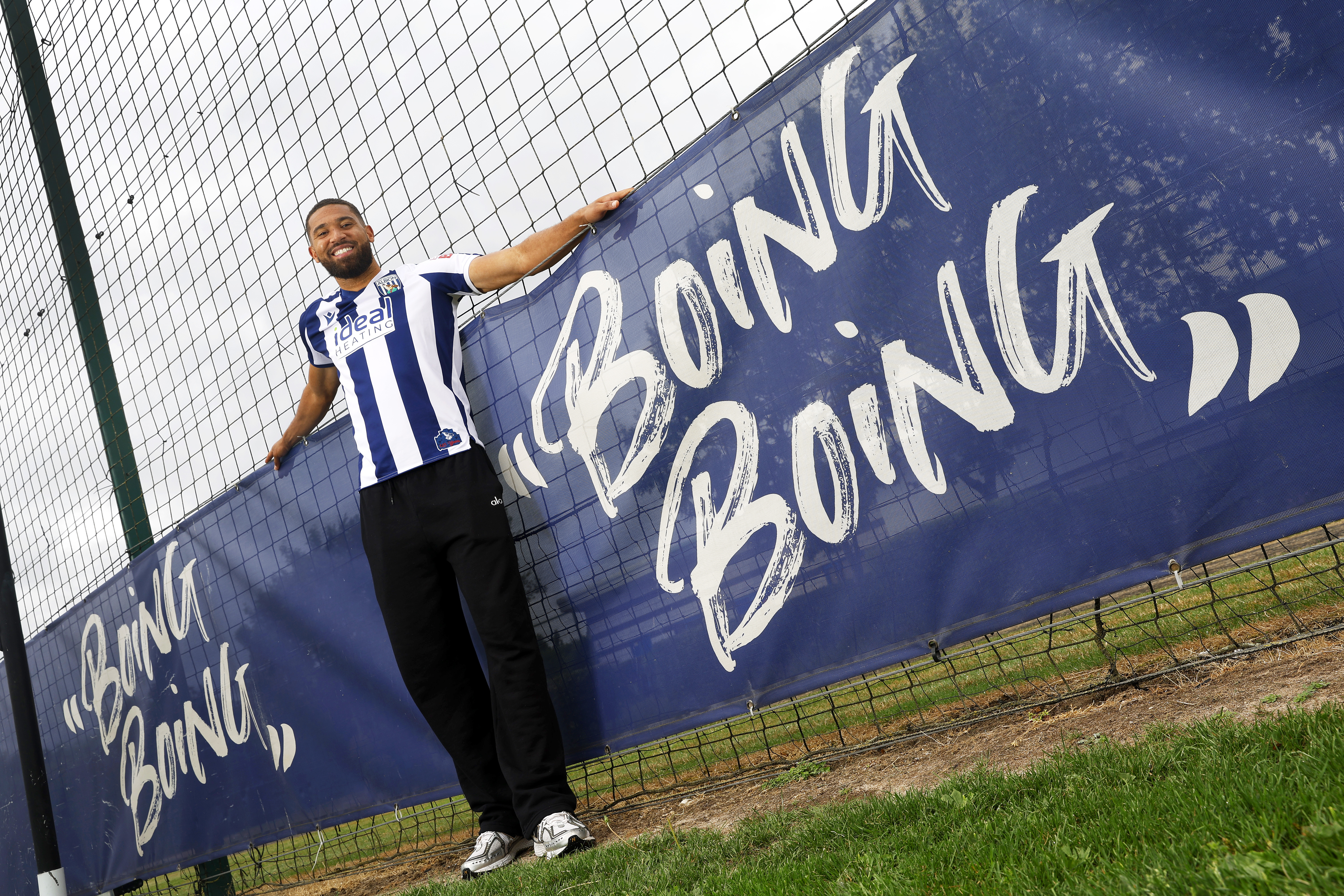 George Campbell stood smiling at the camera in a home WBA shirt next to branding on the pitch which reads 'boing boing'