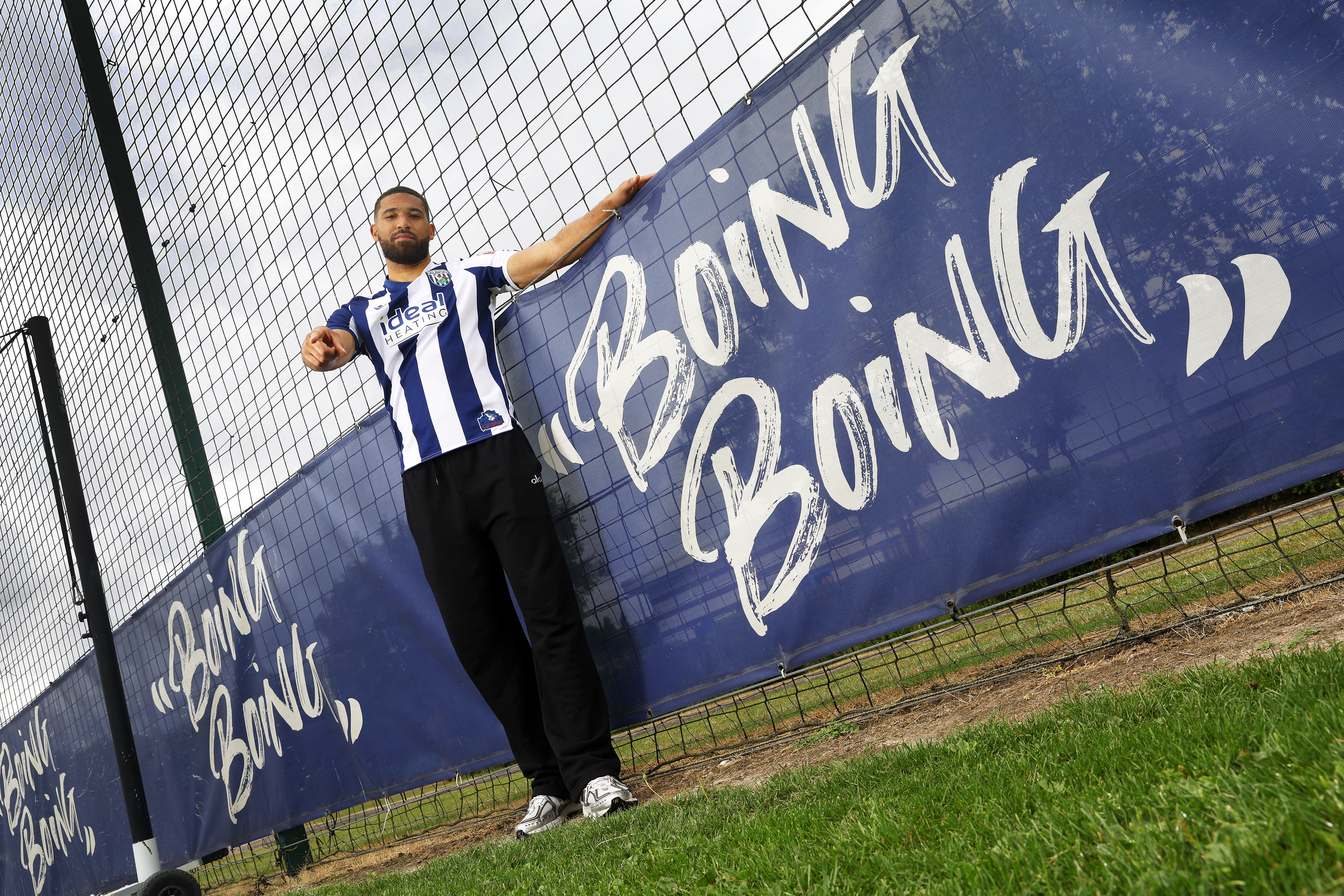 George Campbell stood smiling and pointing at the camera in a home WBA shirt next to branding on the pitch which reads 'boing boing'