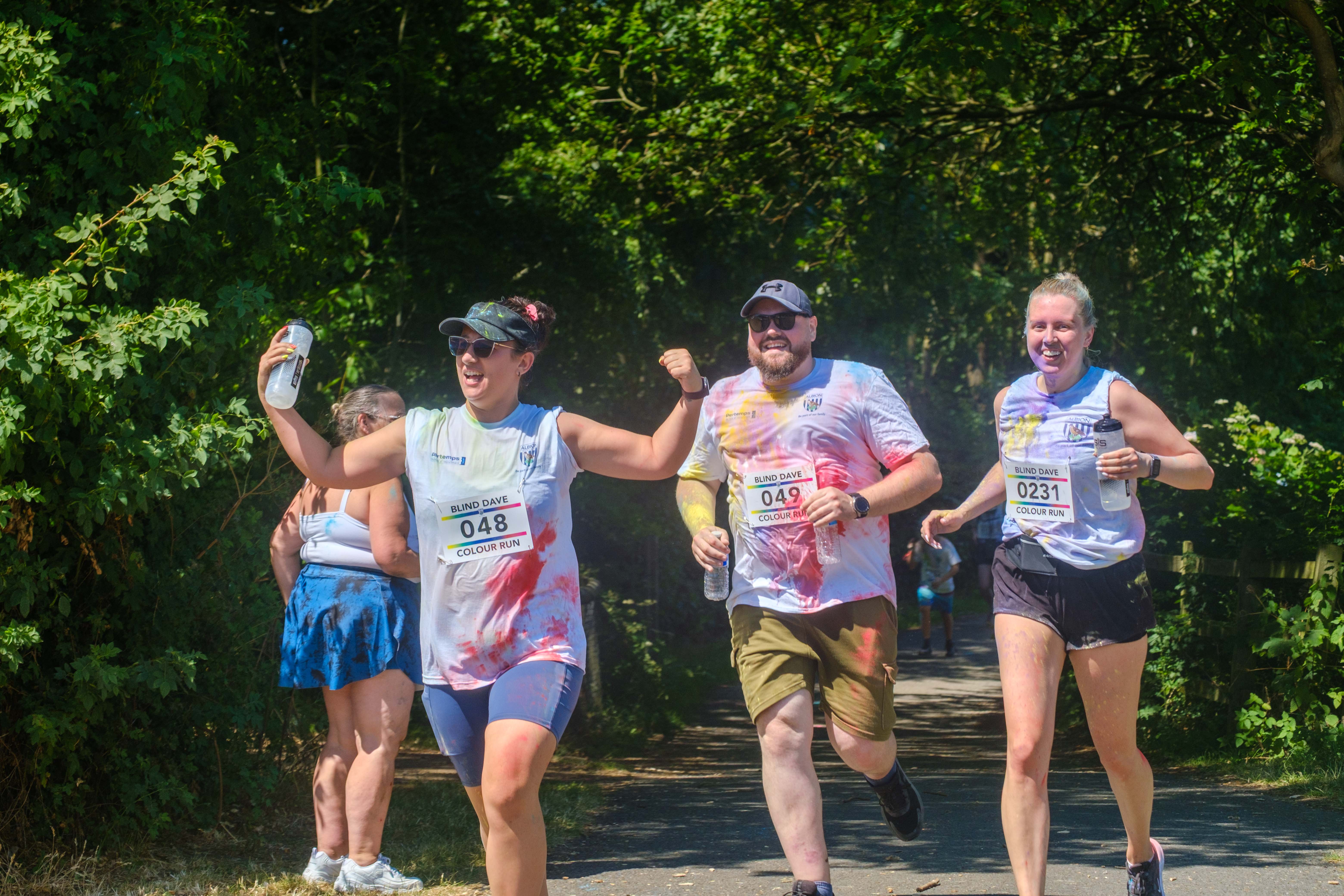 Three runners holding water bottles, 048 flexing her muscles, heading towards the final stretch.