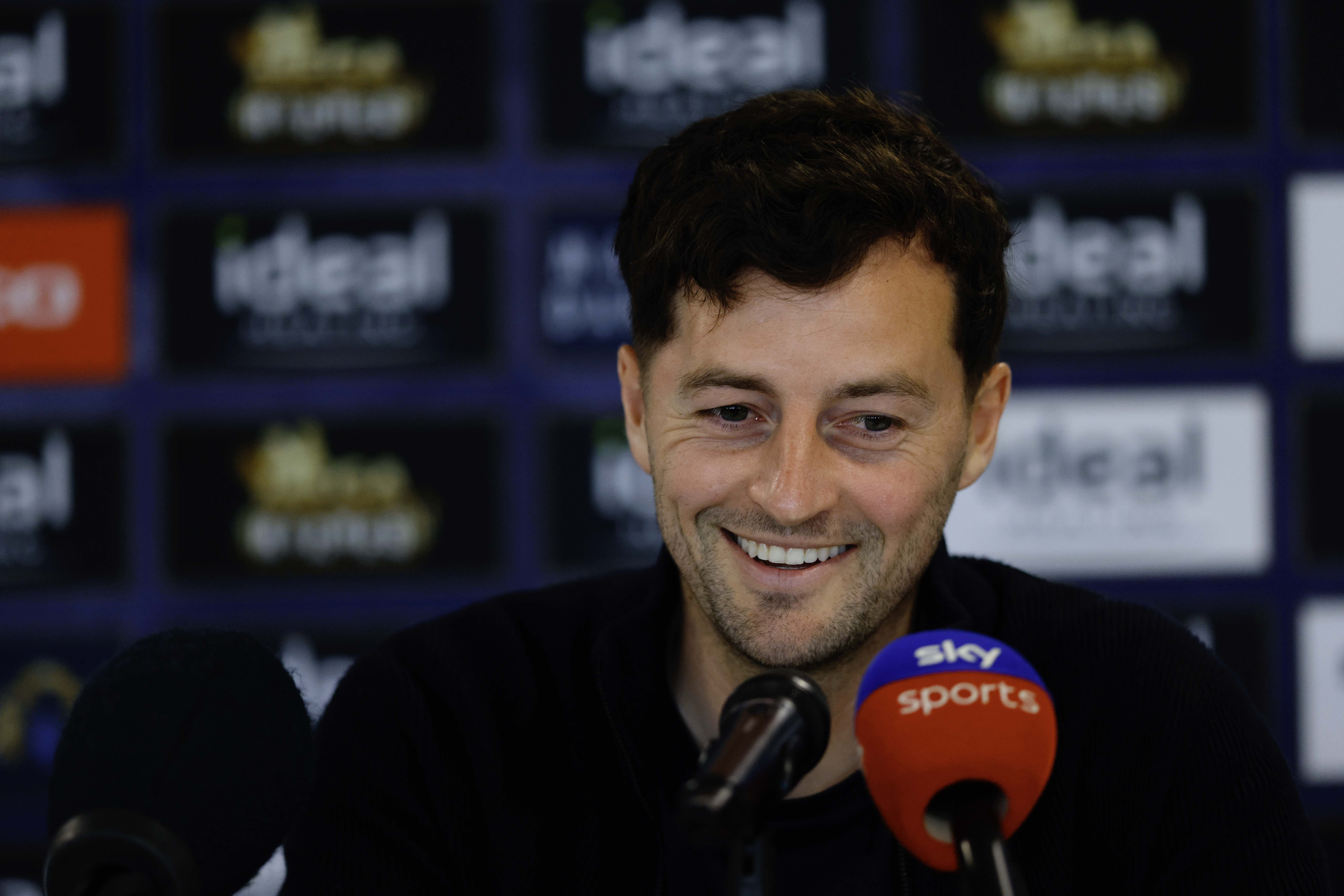 Ryan Mason smiling while talking to the media in front of a sponsor board and behind microphones