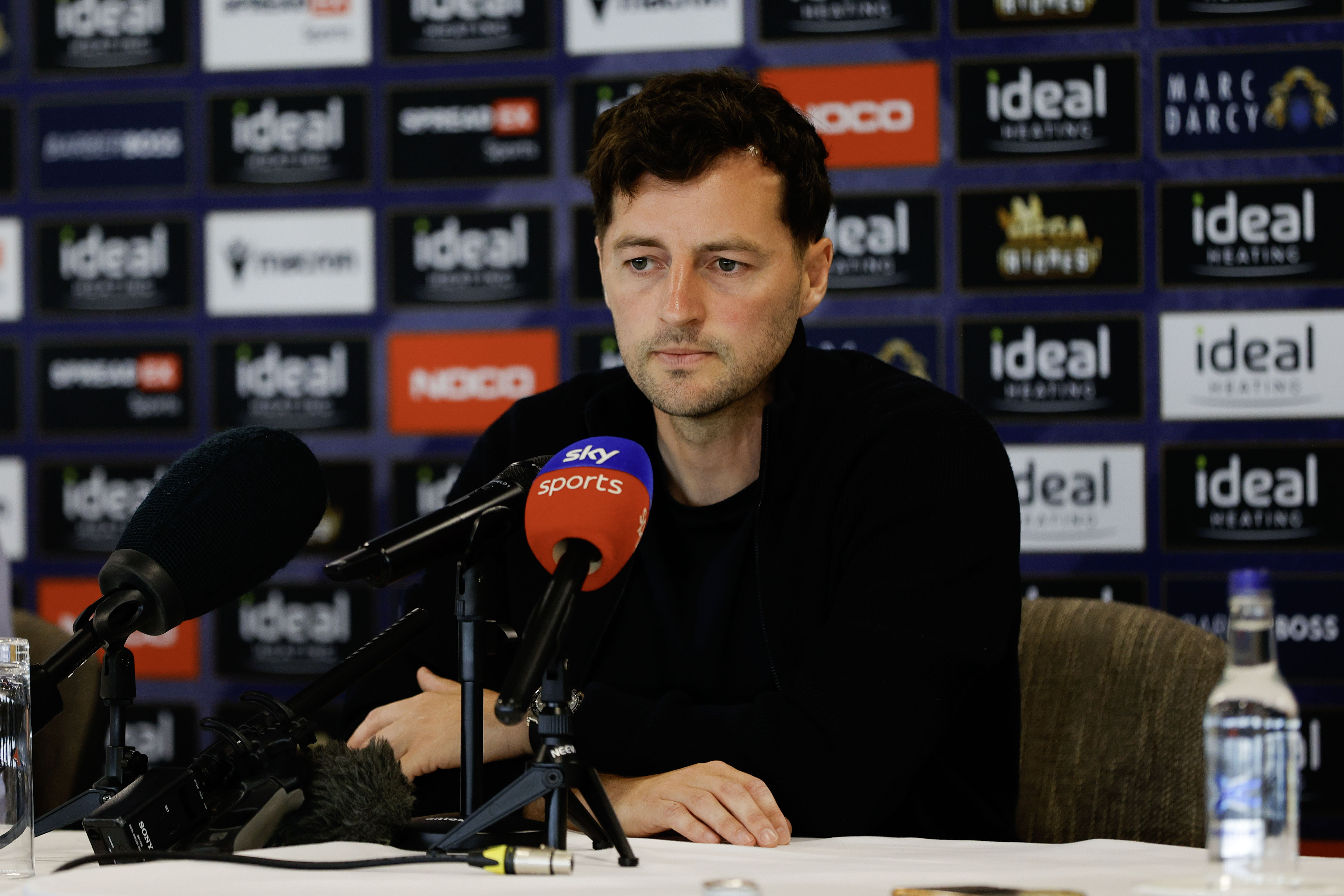 Ryan Mason talking to members of the media in front of a press conference sponsor board and behind a number of microphones