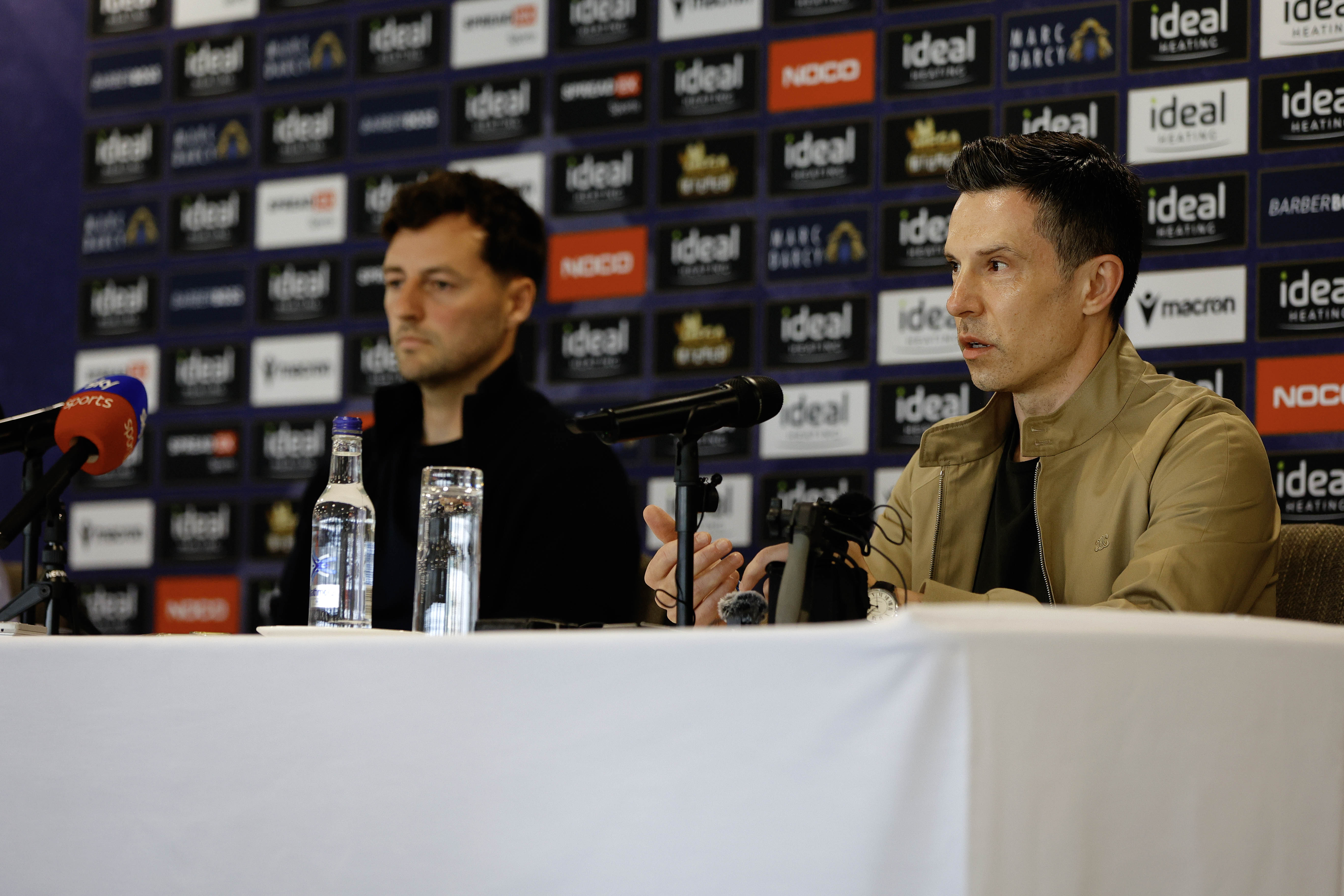 Ryan Mason and Andrew Nestor talking to members of the media in front of a press conference sponsor board and behind a number of microphones
