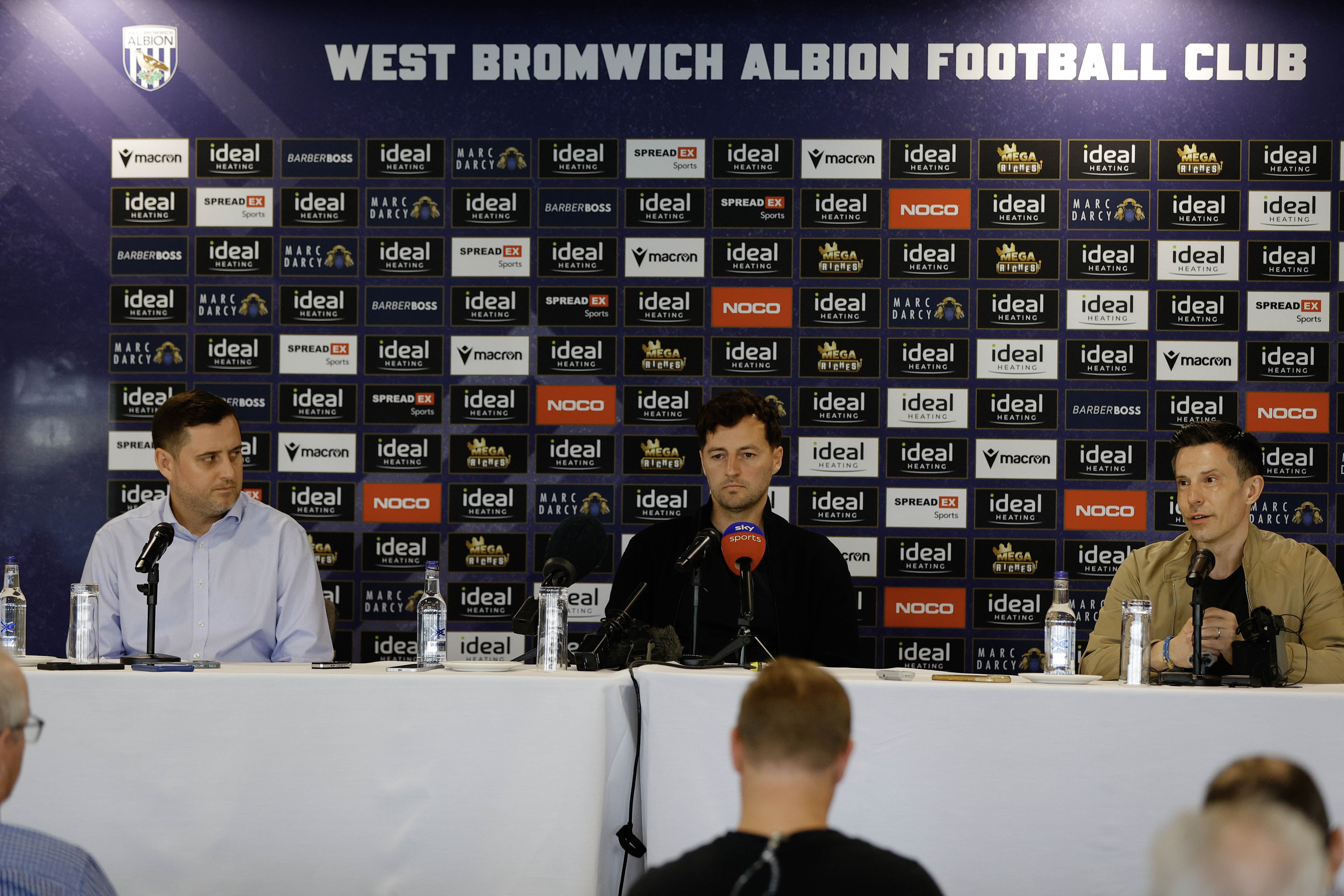 Ryan Mason and Andrew Nestor talking to members of the media in front of a press conference sponsor board and behind a number of microphones