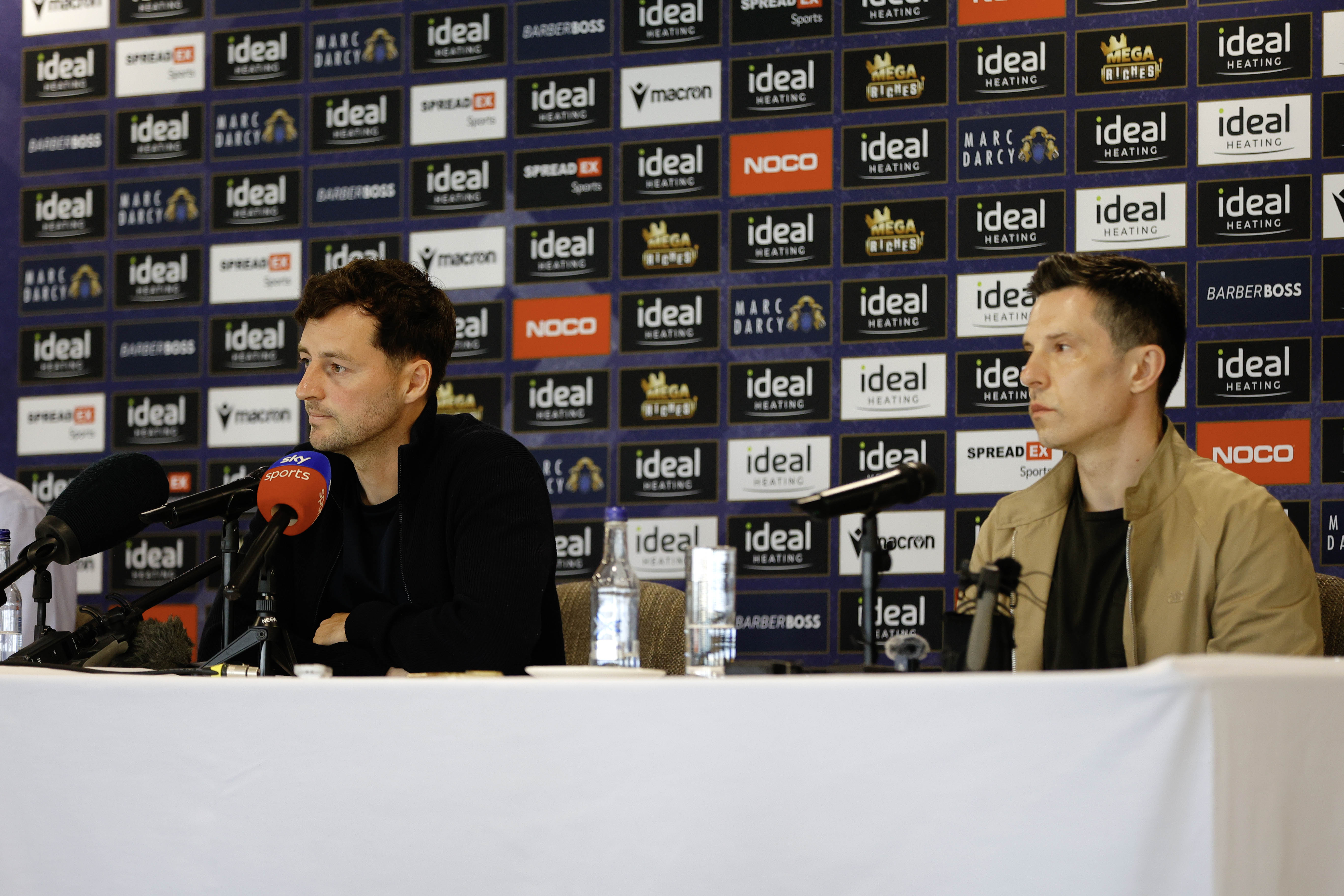 Ryan Mason and Andrew Nestor talking to members of the media in front of a press conference sponsor board and behind a number of microphones