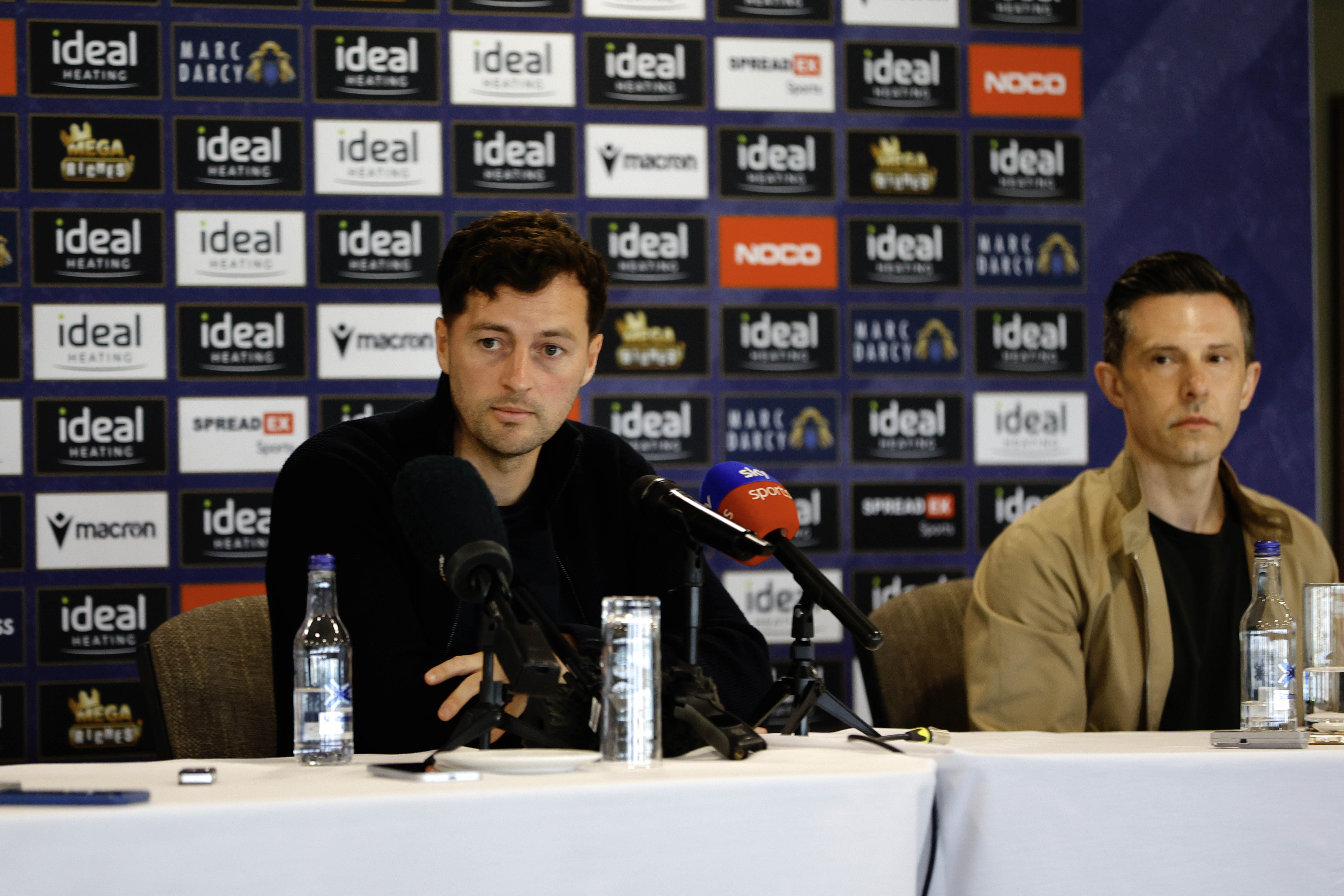 Ryan Mason and Andrew Nestor talking to members of the media in front of a press conference sponsor board and behind a number of microphones