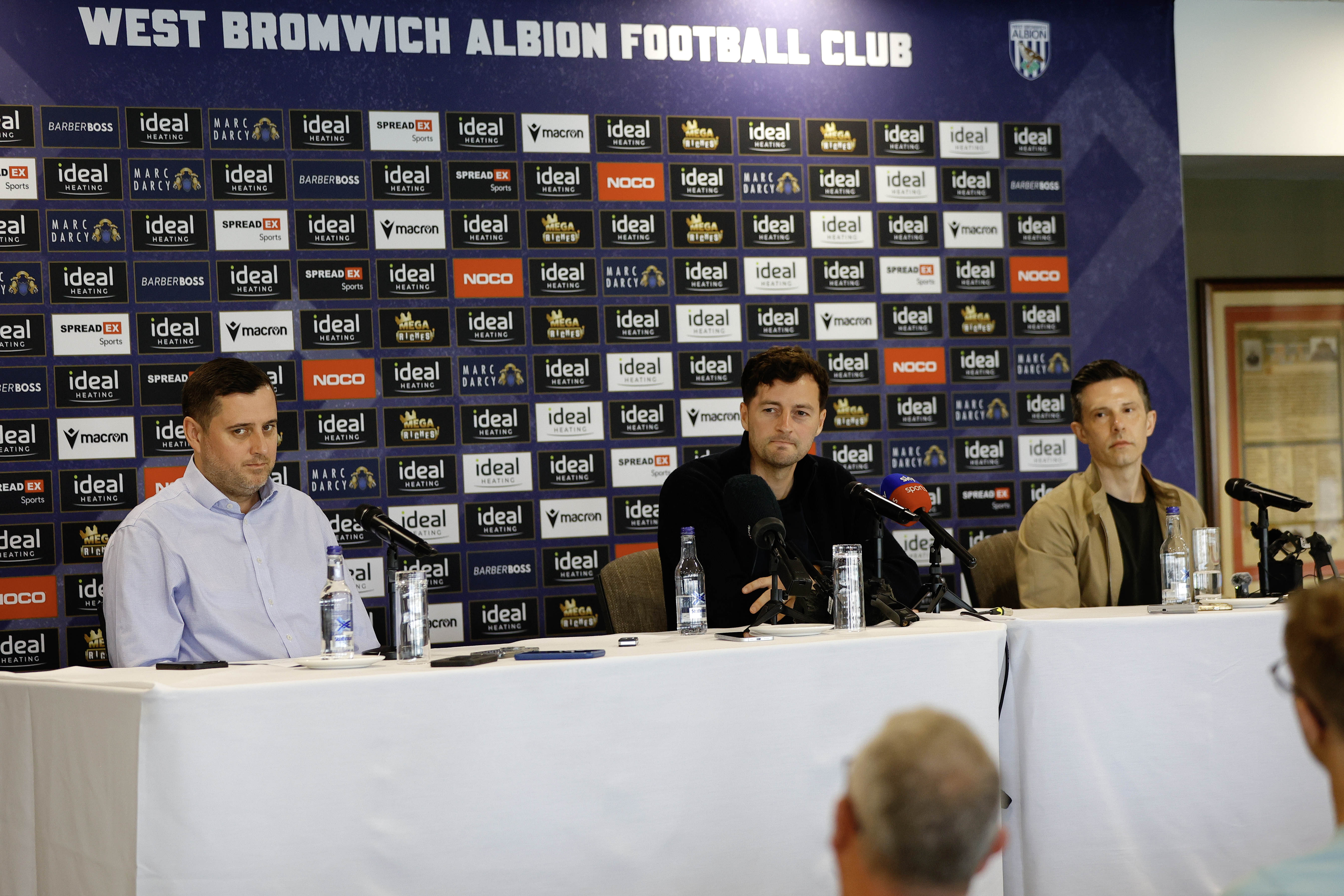 Ryan Mason and Andrew Nestor talking to members of the media in front of a press conference sponsor board and behind a number of microphones
