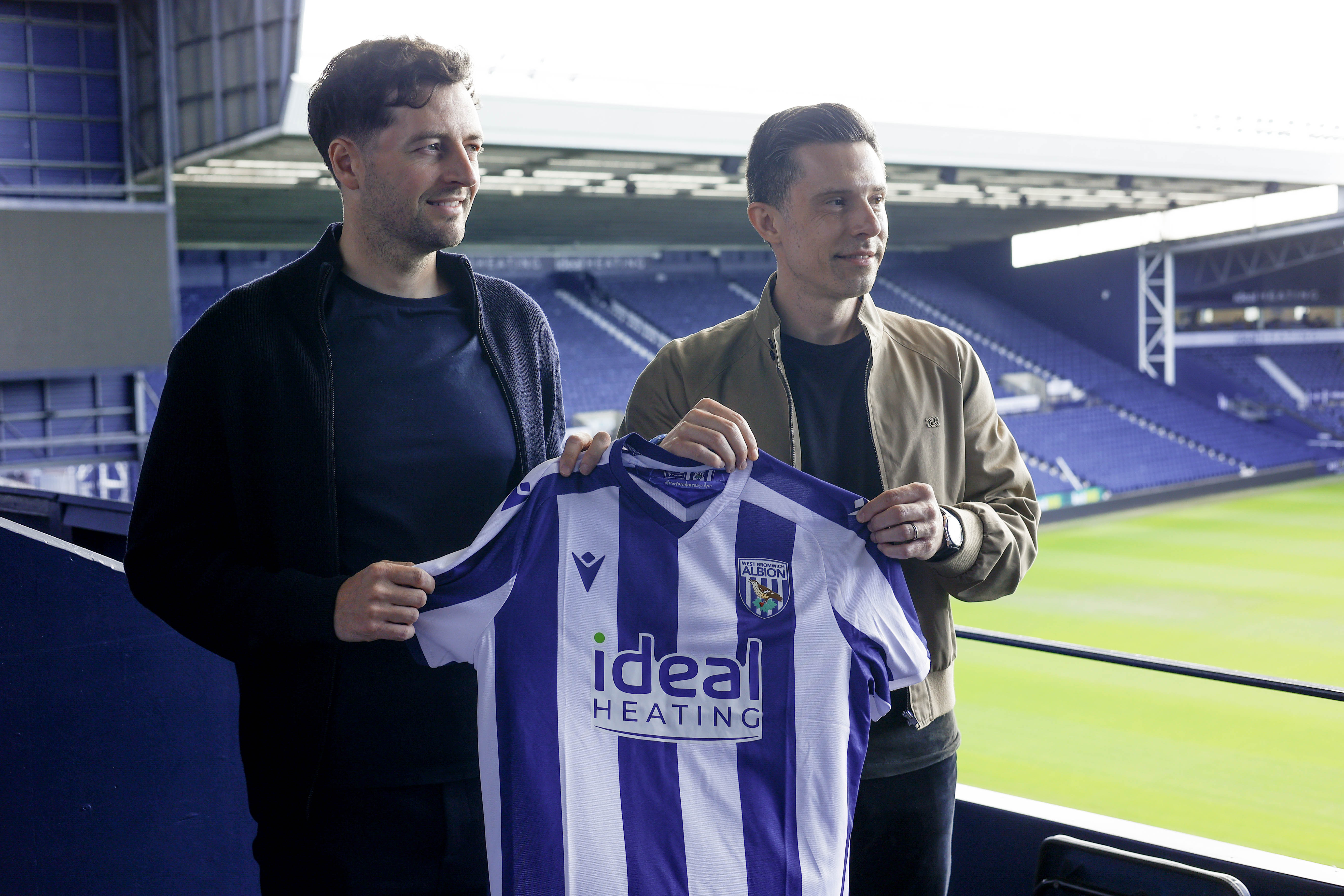 Ryan Mason and Andrew Nestor posing for a photo while holding up a home WBA shirt in front of the West Stand at The Hawthorns