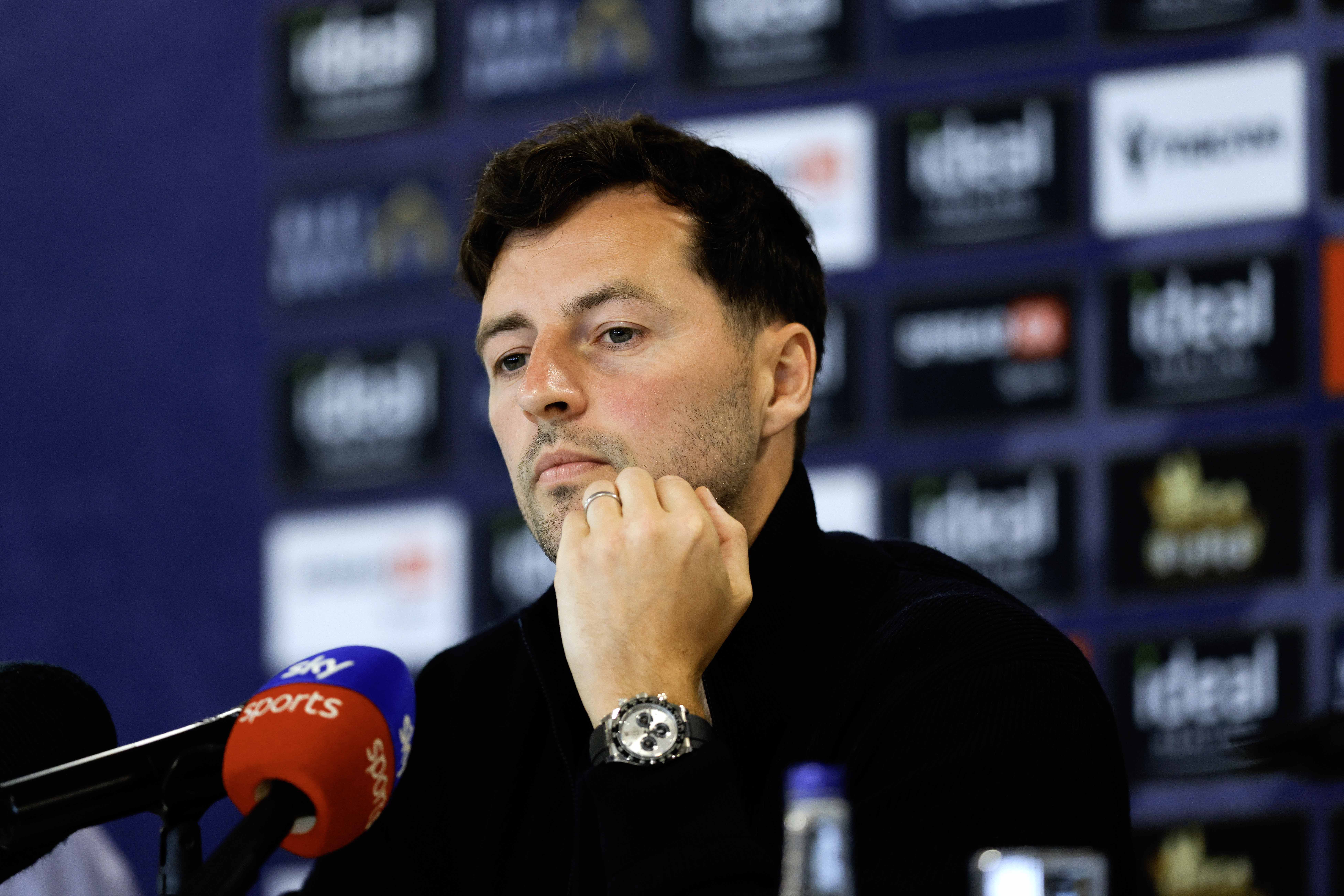 Ryan Mason talking to members of the media in front of a press conference sponsor board and behind a number of microphones
