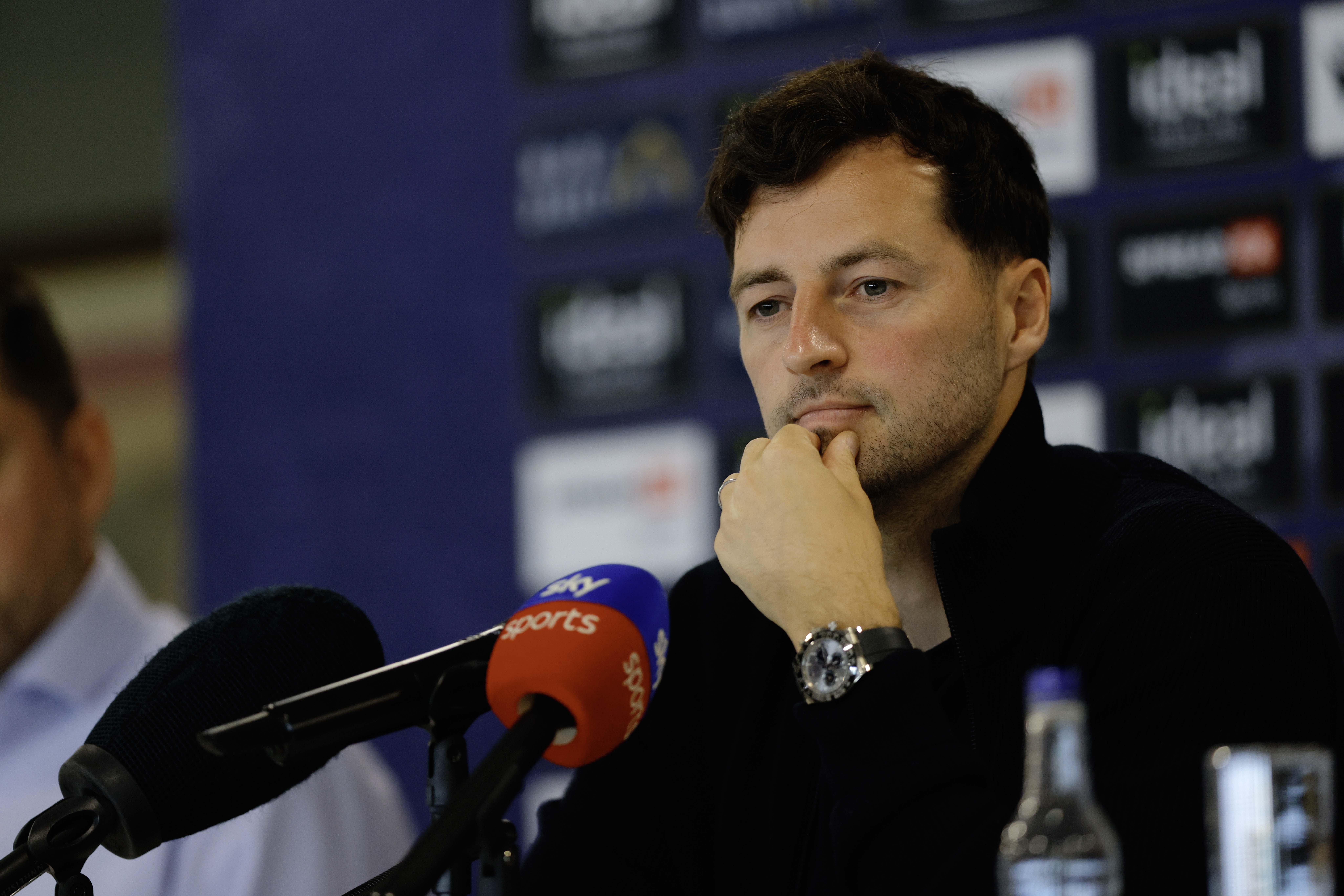 Ryan Mason talking to members of the media in front of a press conference sponsor board and behind a number of microphones