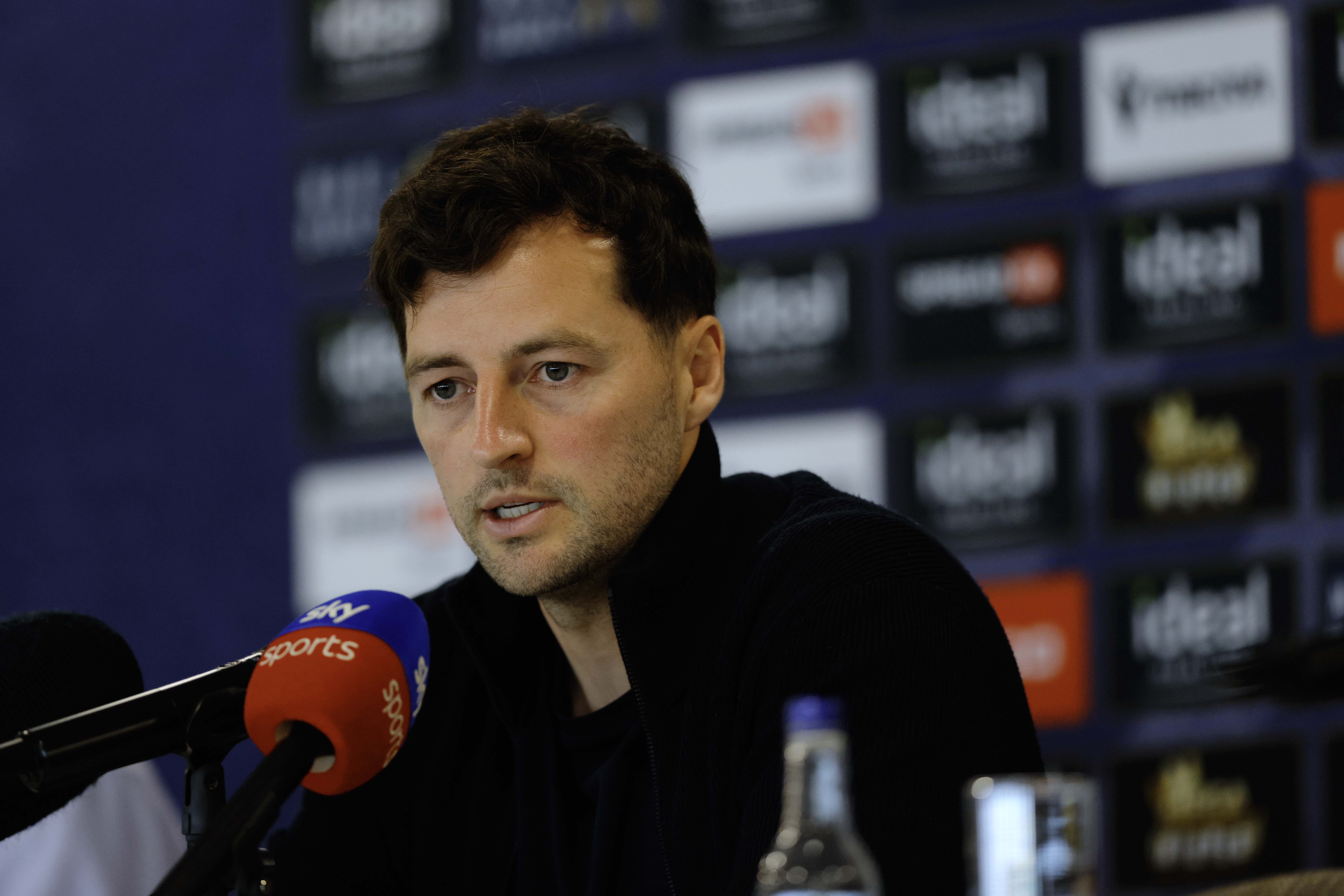 Ryan Mason talking to members of the media in front of a press conference sponsor board and behind a number of microphones