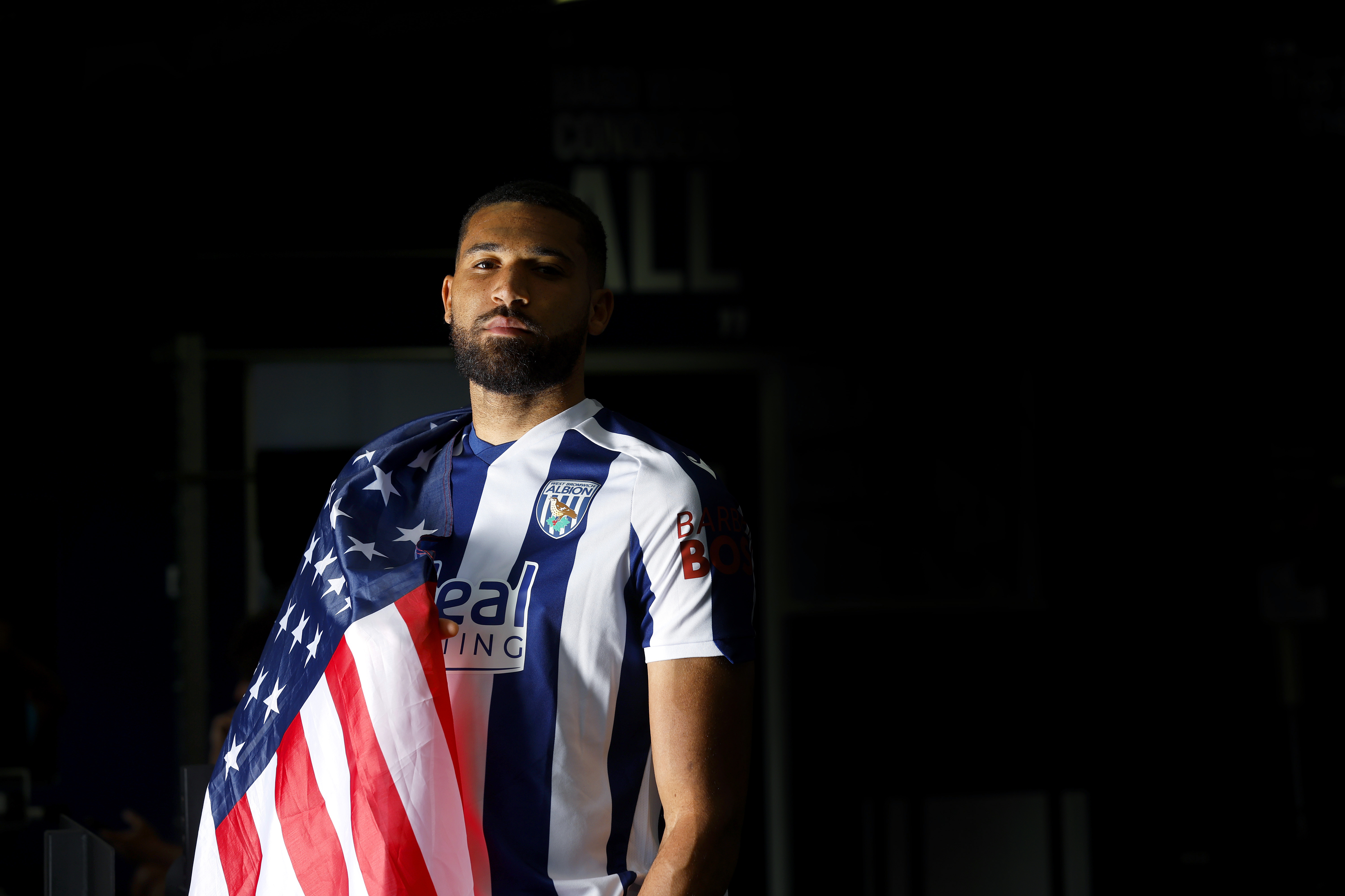 George Campbell looking at the camera while stood in a home WBA shirt with an American flag wrapped round his shoulder