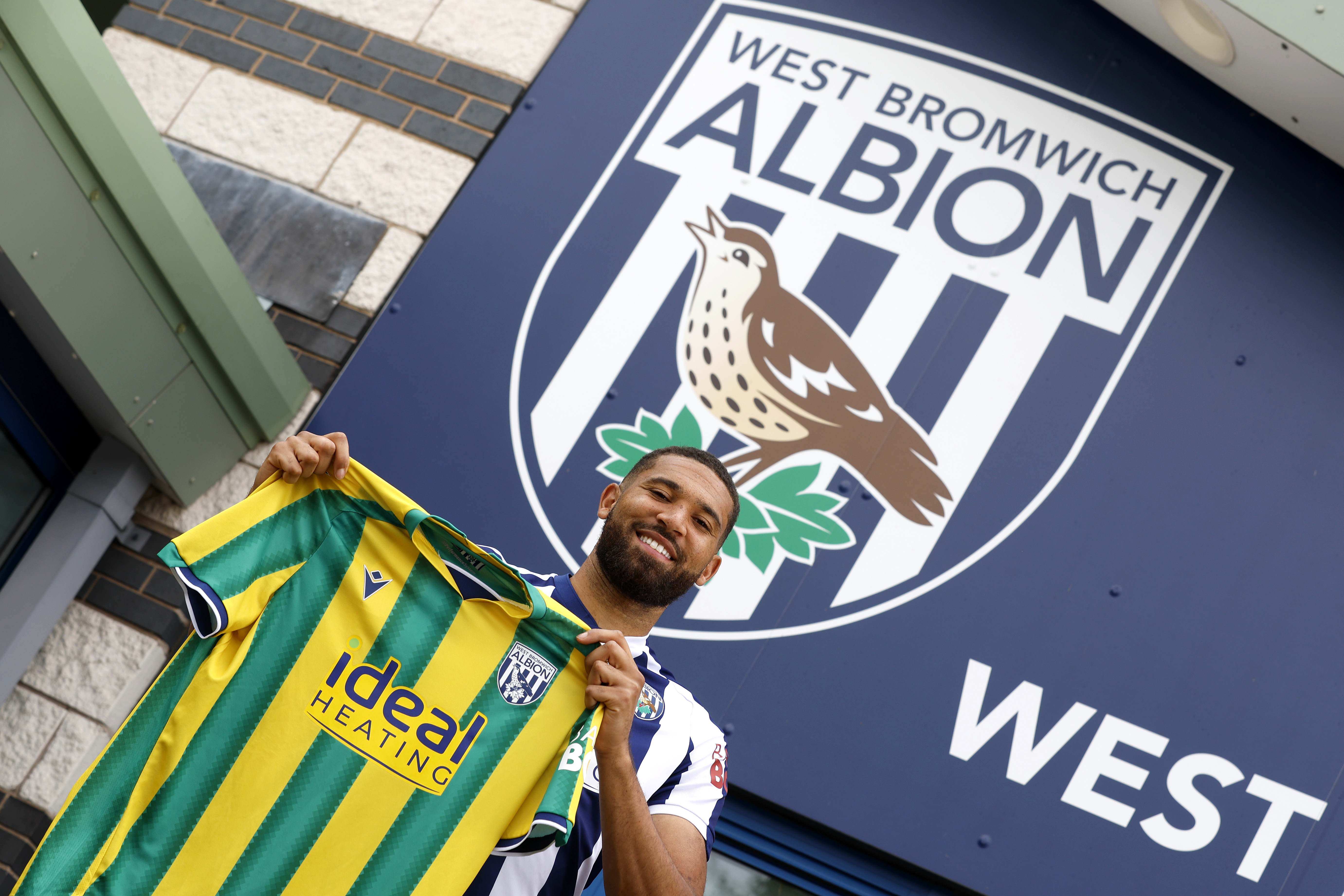 George Campbell smiling at the camera while holding up an away WBA shirt in front of a big WBA badge 