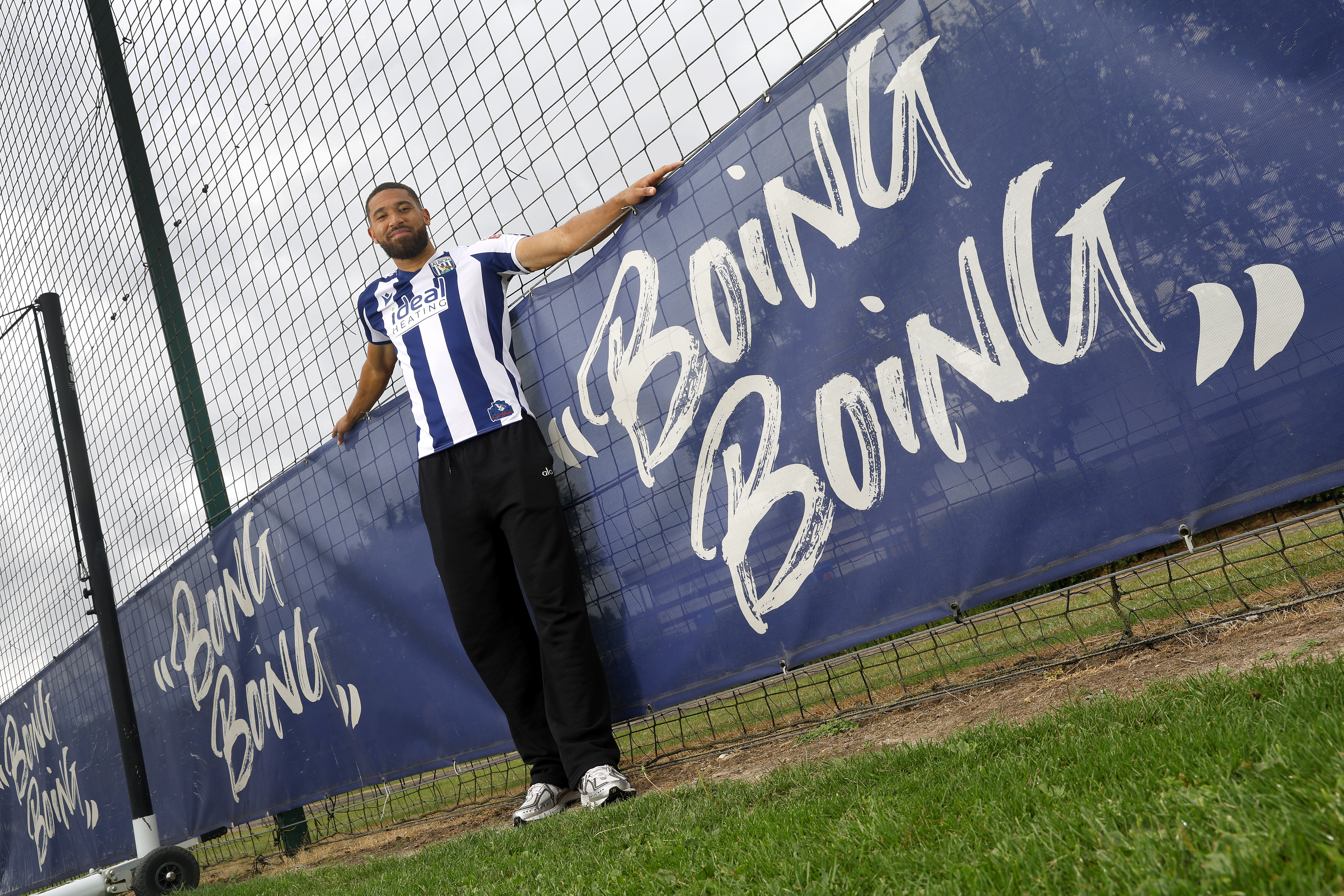 George Campbell smiling at the camera while wearing a home WBA shirt stood next to branding round the training pitch which reads 'boing boing'