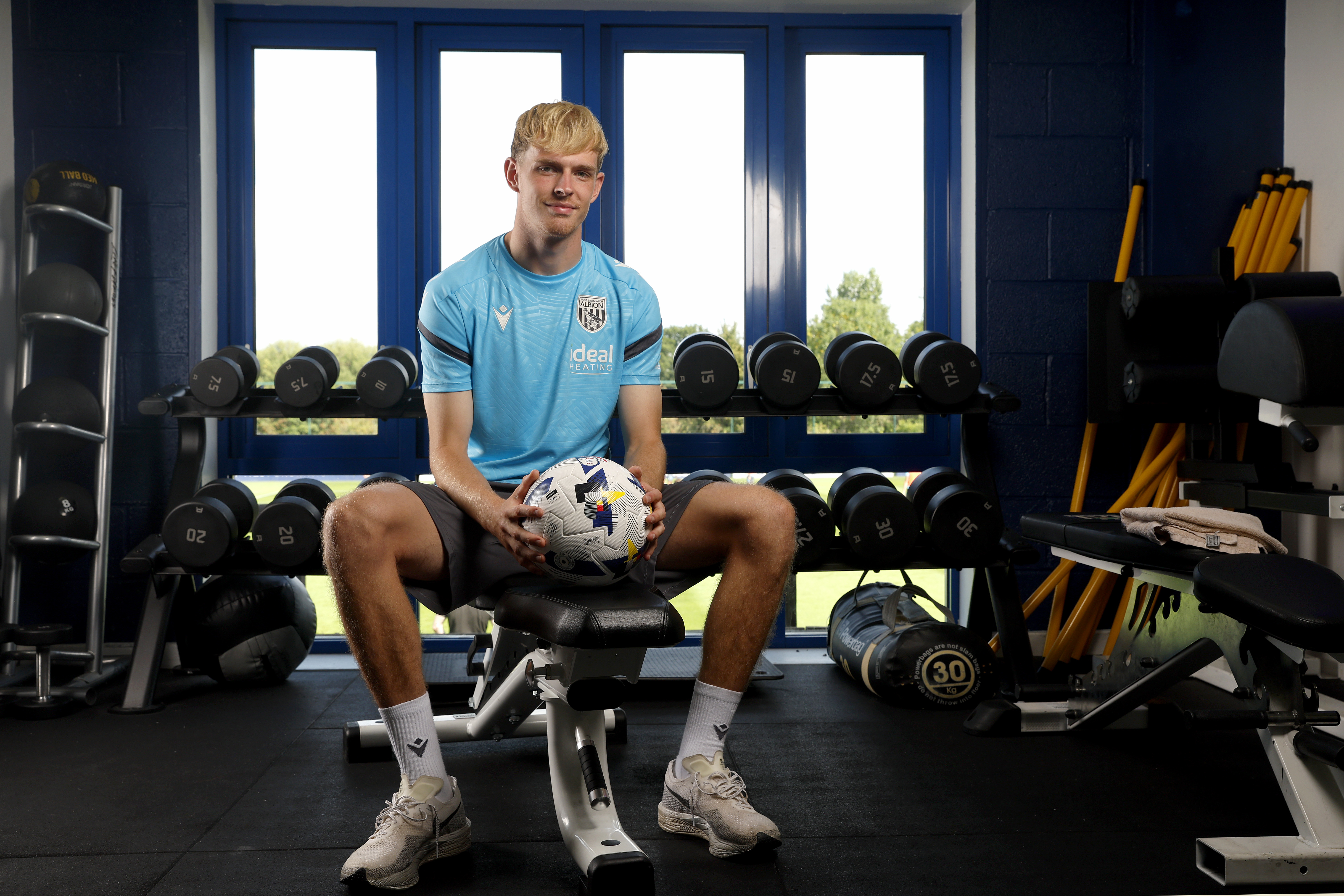 Toby Collyer sat on a bench in the gym smiling at the camera holding a ball