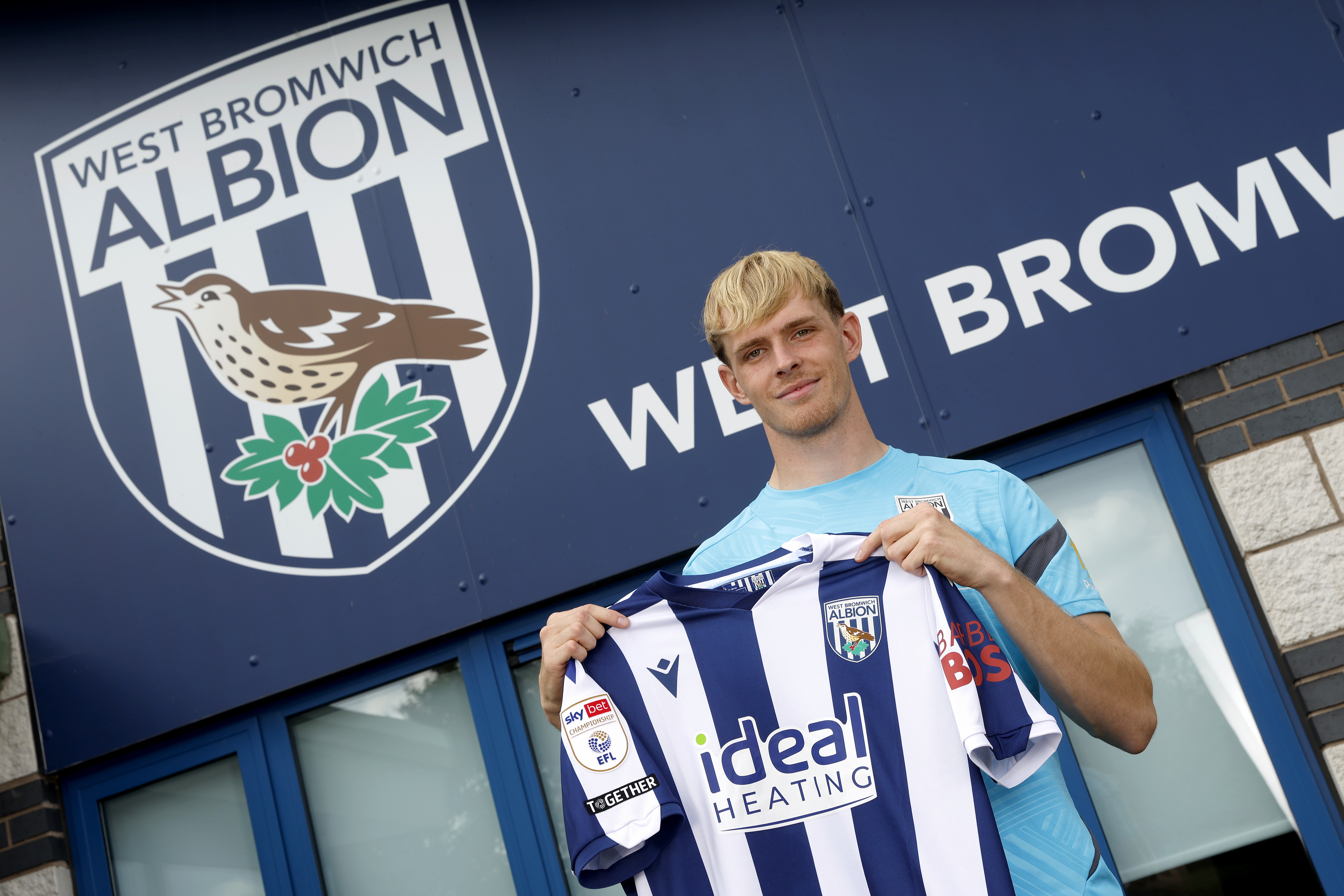 Toby Collyer smiling and looking at the camera while stood outside the training ground holding up a WBA shirt 