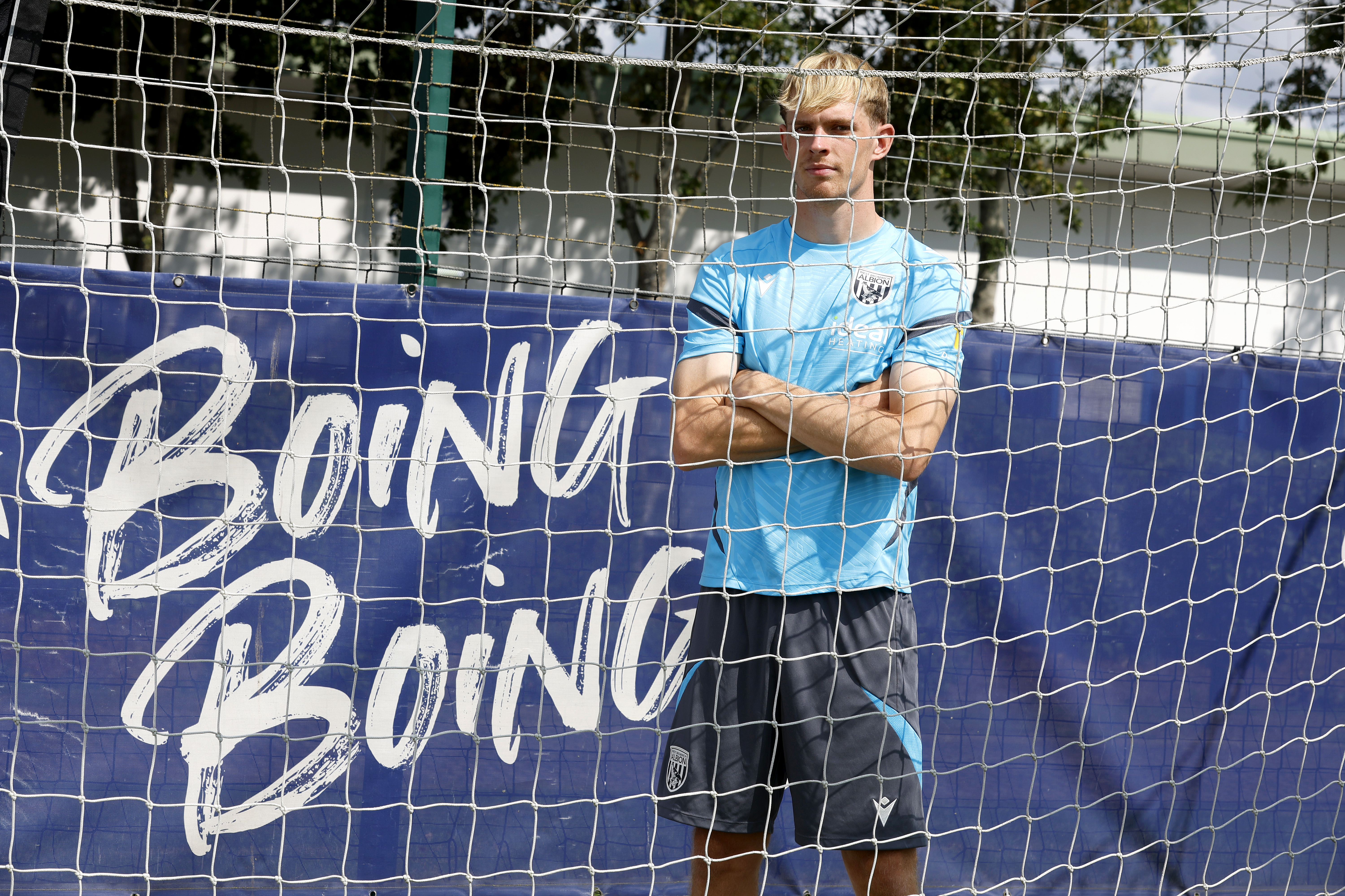 Toby Collyer looking through a goal net with his arms crossed next to branding saying "boing boing'