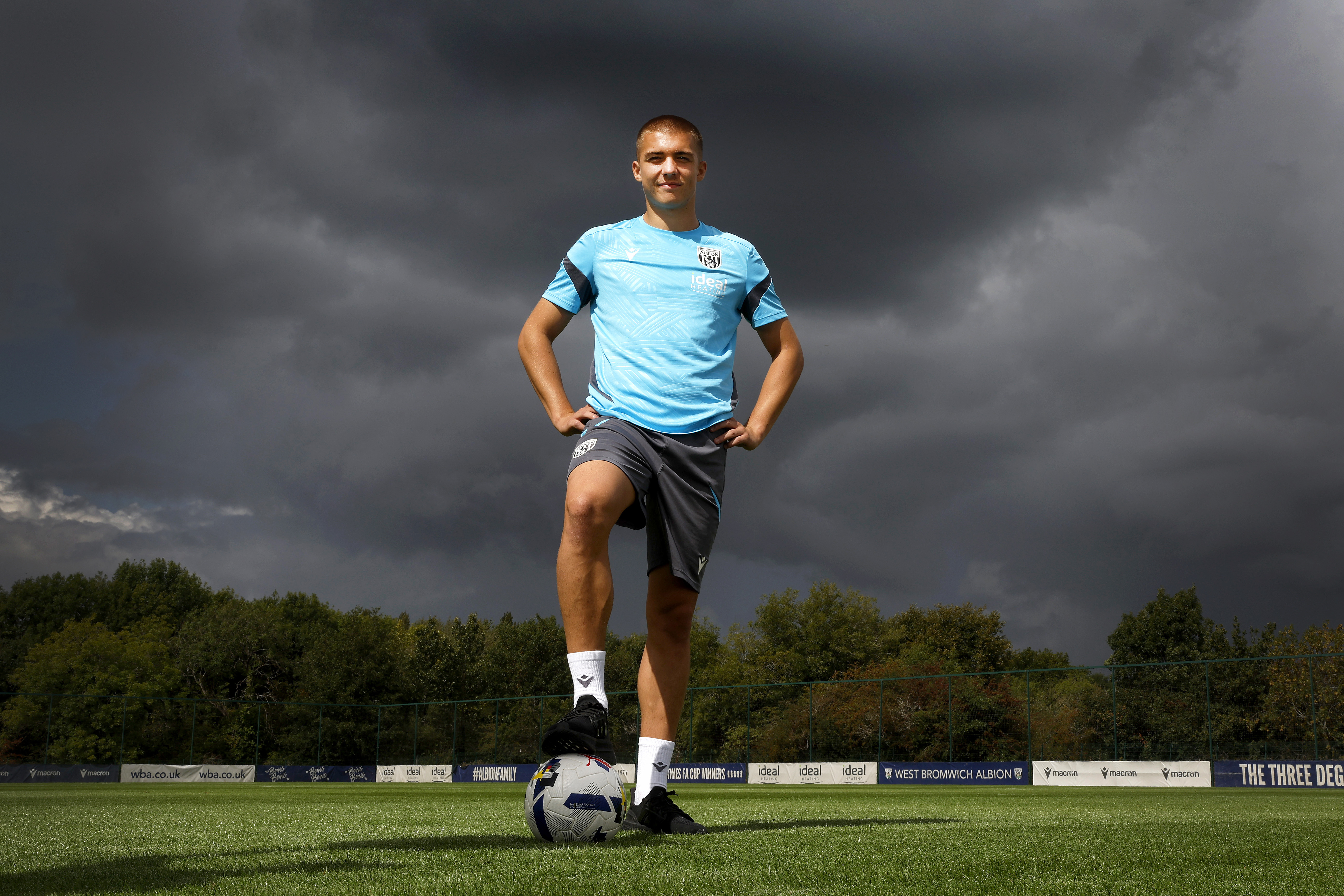 Alfie Gilchrist stood with his foot on a ball and his hands on his hips out on a training pitch smiling at the camera 