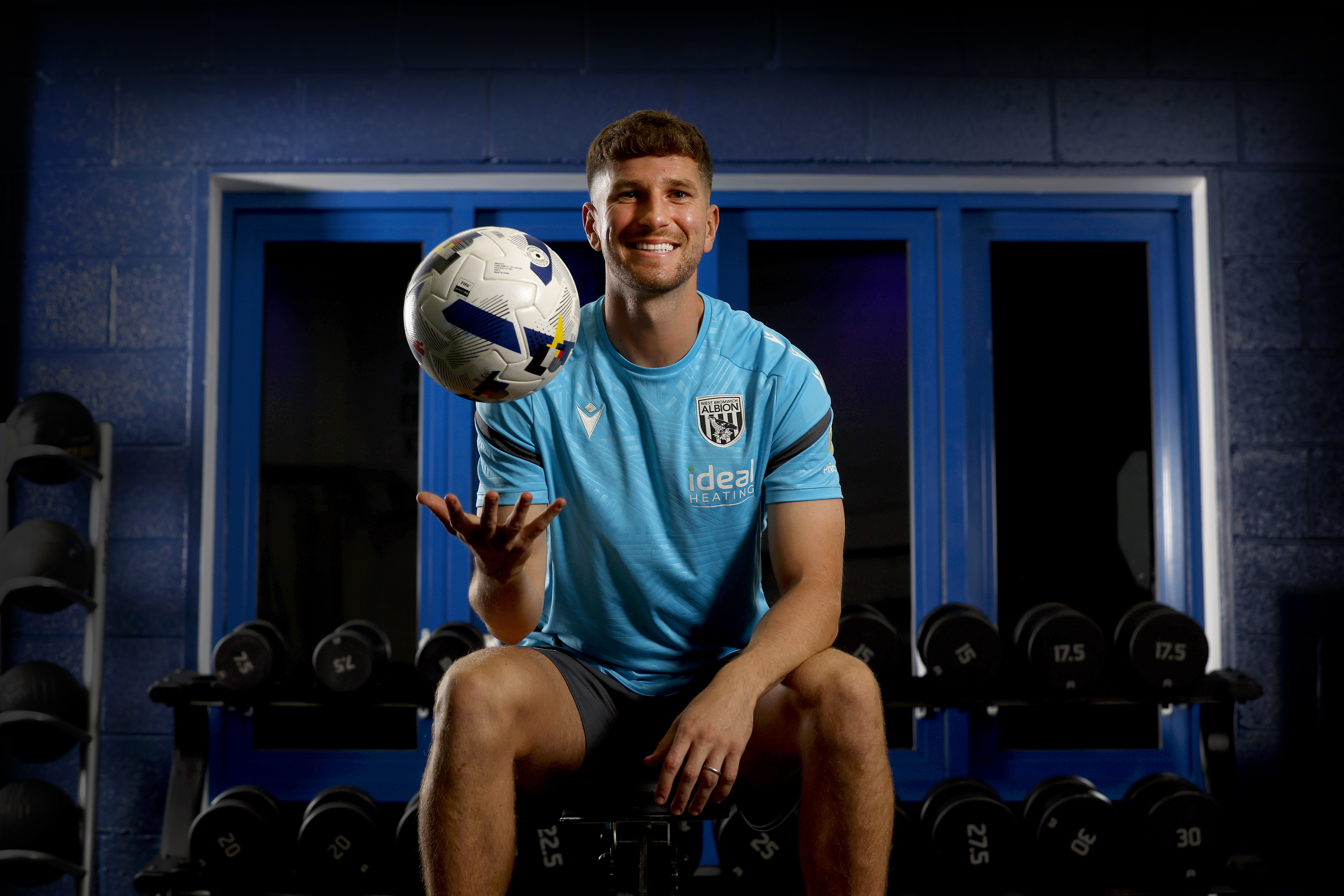 Chris Mepham smiling at the camera while holding a ball and being sat down in the gym