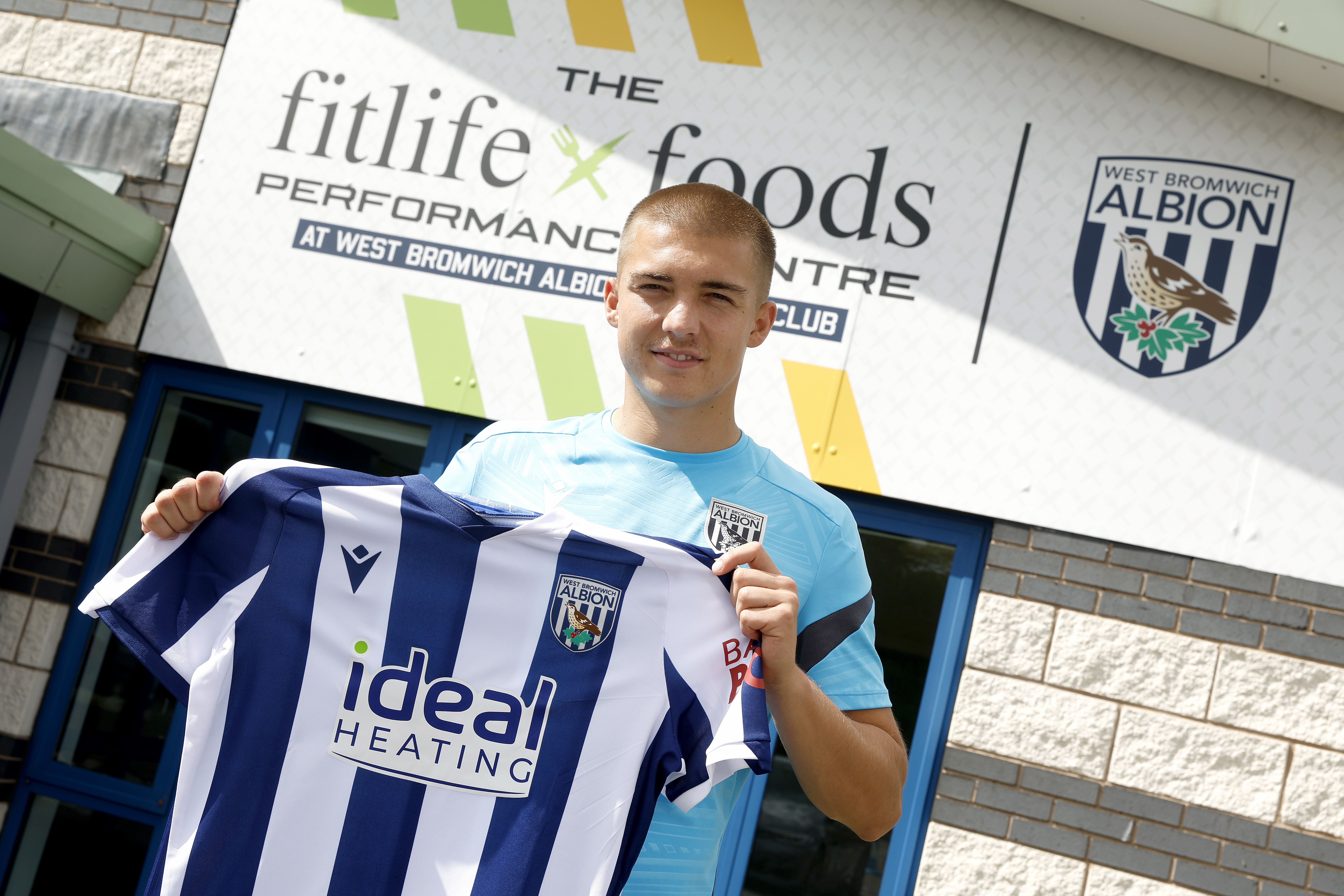 Alfie Gilchrist smiling at the camera while holding up a home WBA shirt stood outside the training ground