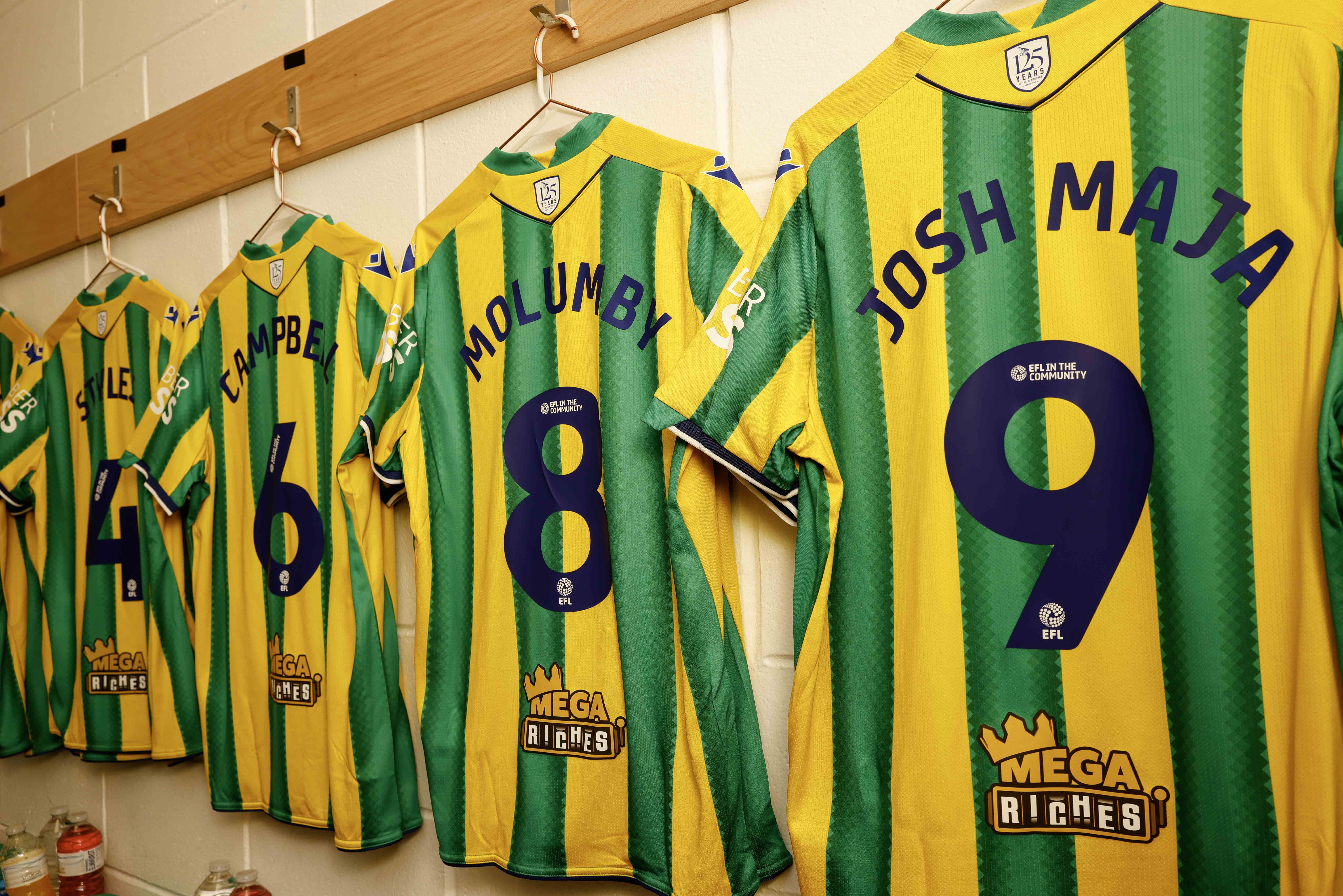 Four green and yellow away shirts hanging up in a dressing room with names and numbers on the back