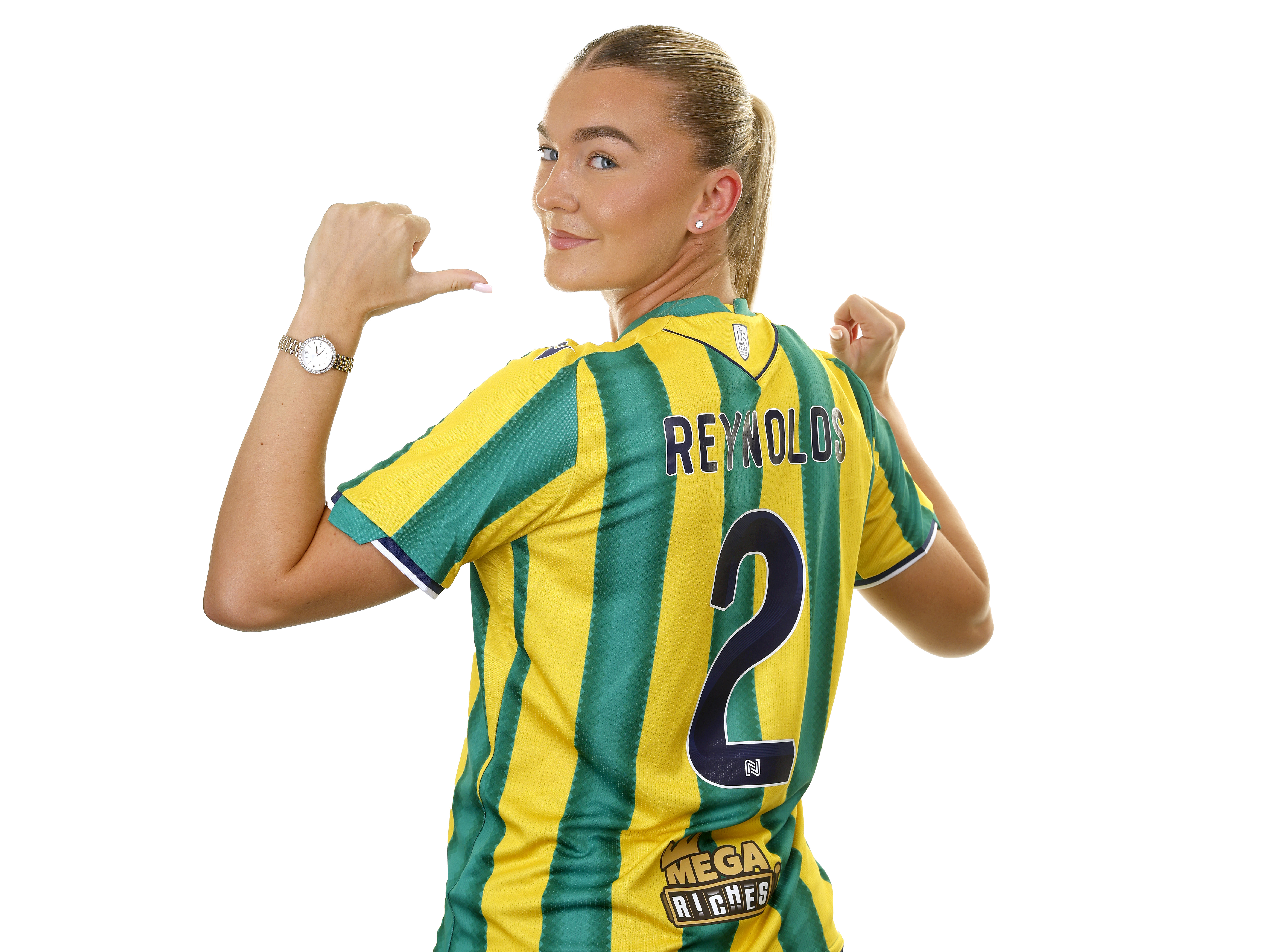 Taylor Reynolds smiling at the camera in the green and yellow away kit pointing to her name and number on the back of her shirt 