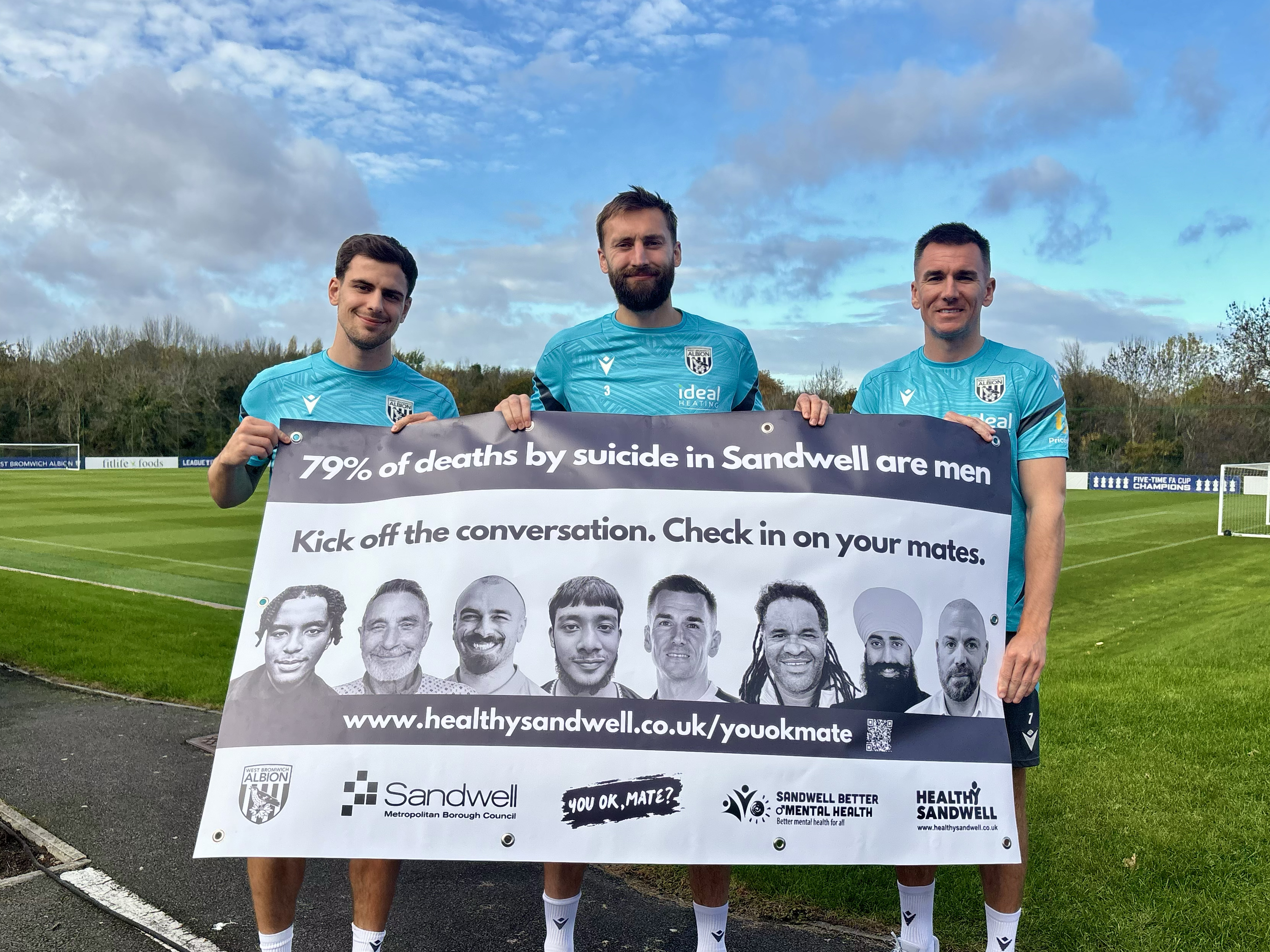 Jayson Molumby, Nat Phillips and Jed Wallace holding up a 'You Ok Mate' banner and smiling at the camera while stood in front of a pitch