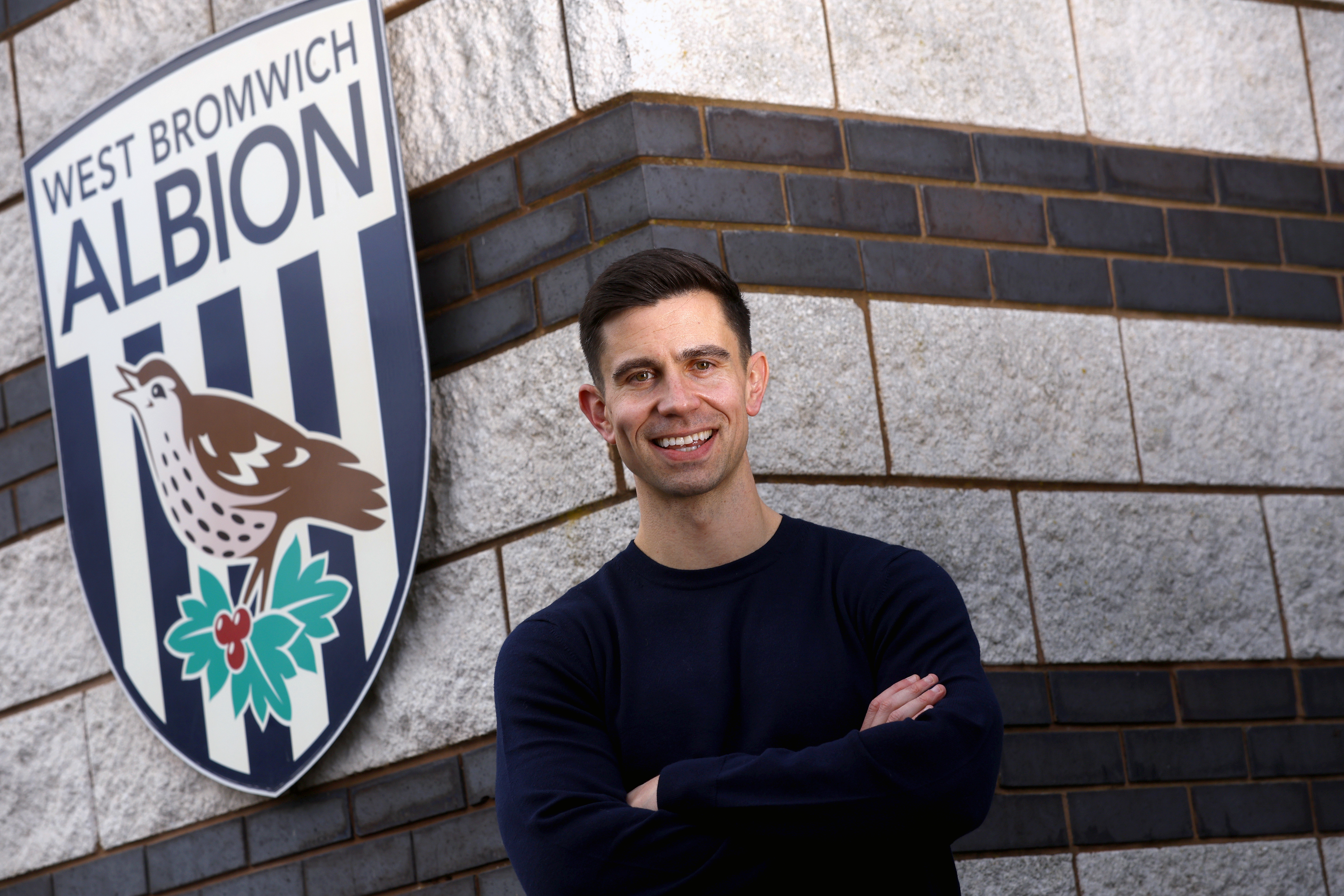 Eric Ramsay posing for a photo by a WBA badge smiling at the camera 