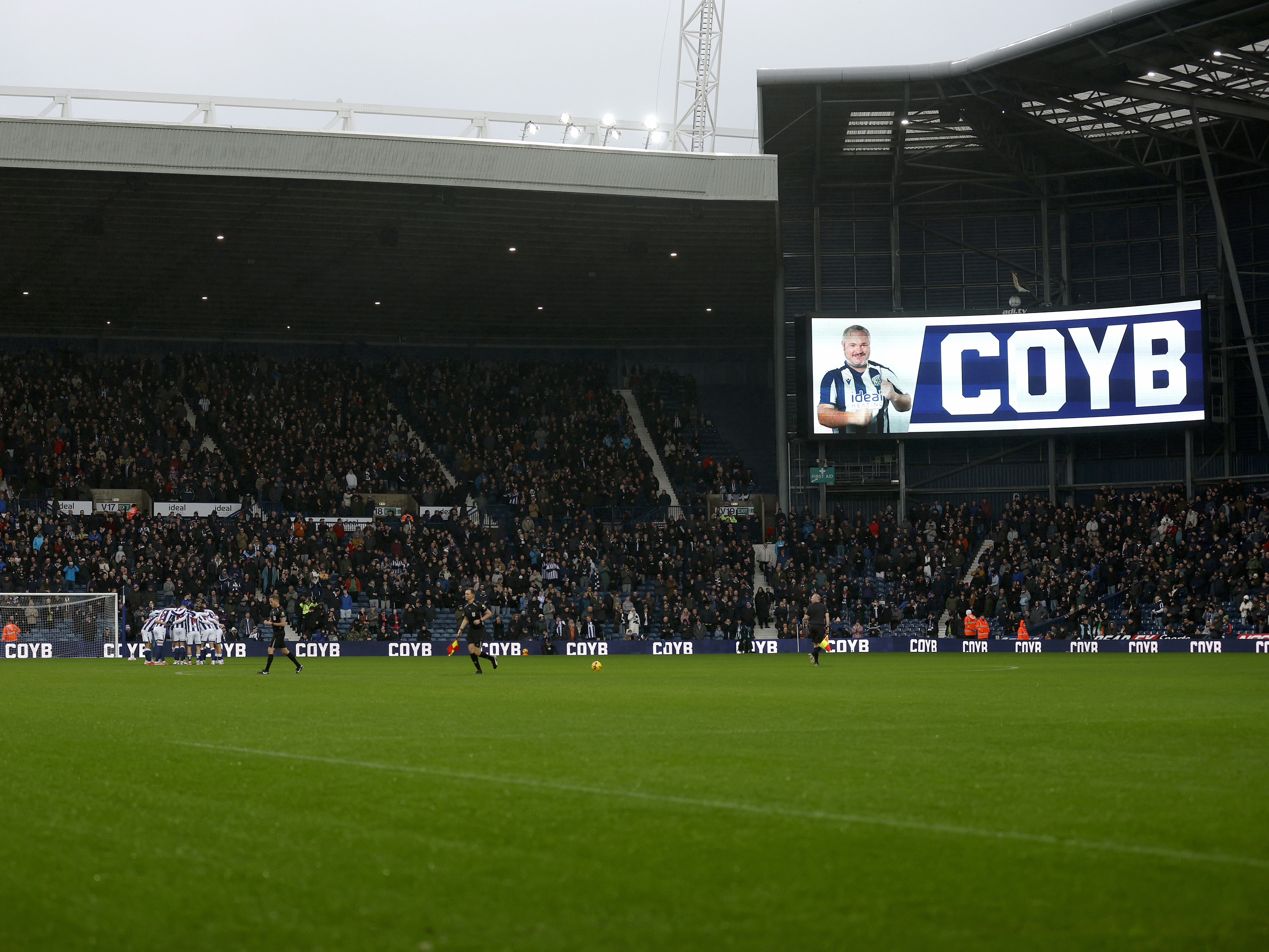 WBA in a pre-match huddle with the Brummie Road End behind them and the big screen displaying a 'COYB' message 