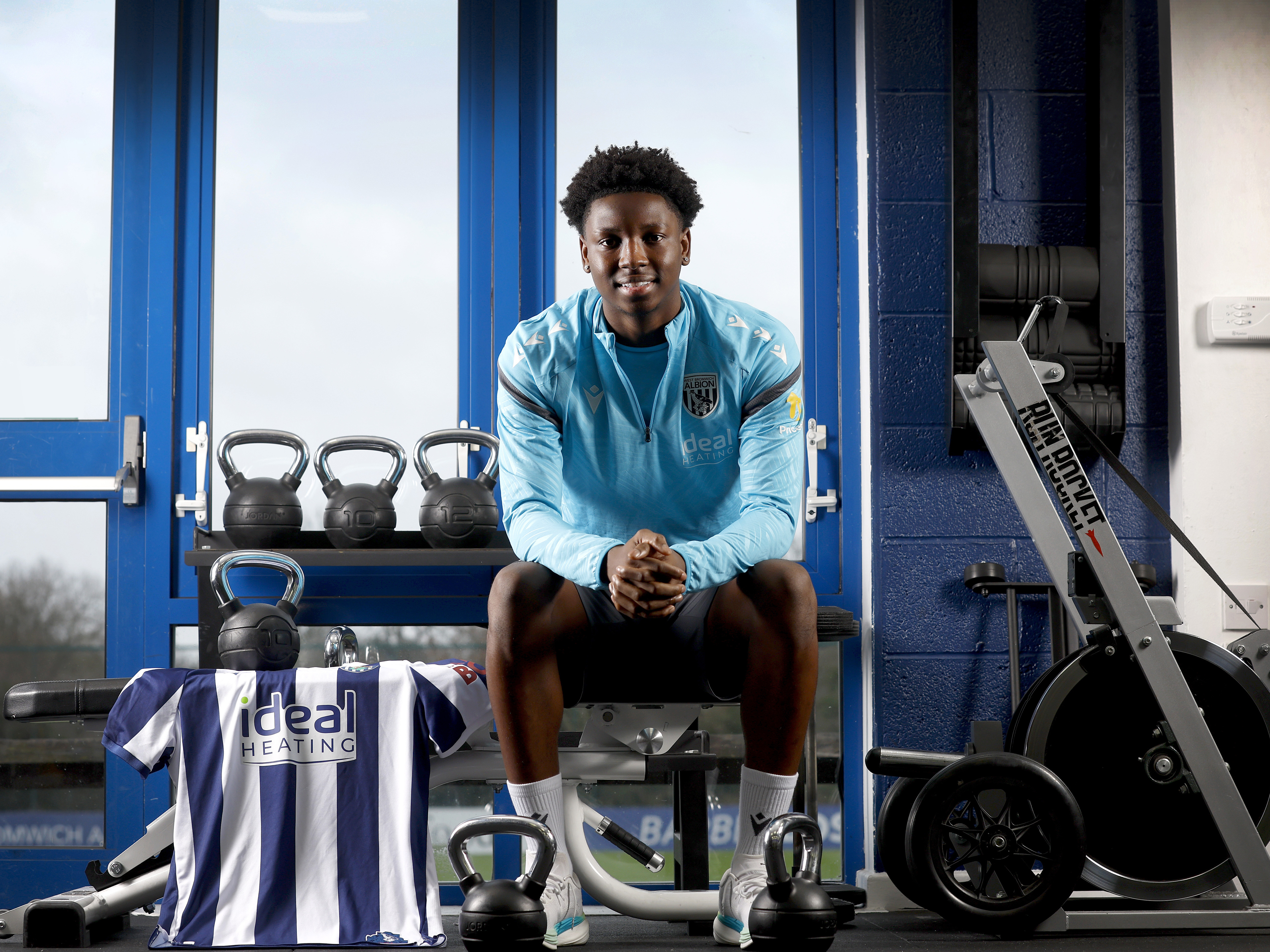 Jamaldeen Jimoh-Aloba smiling at the camera while sat in the gym on a bench with a home shirt next to him
