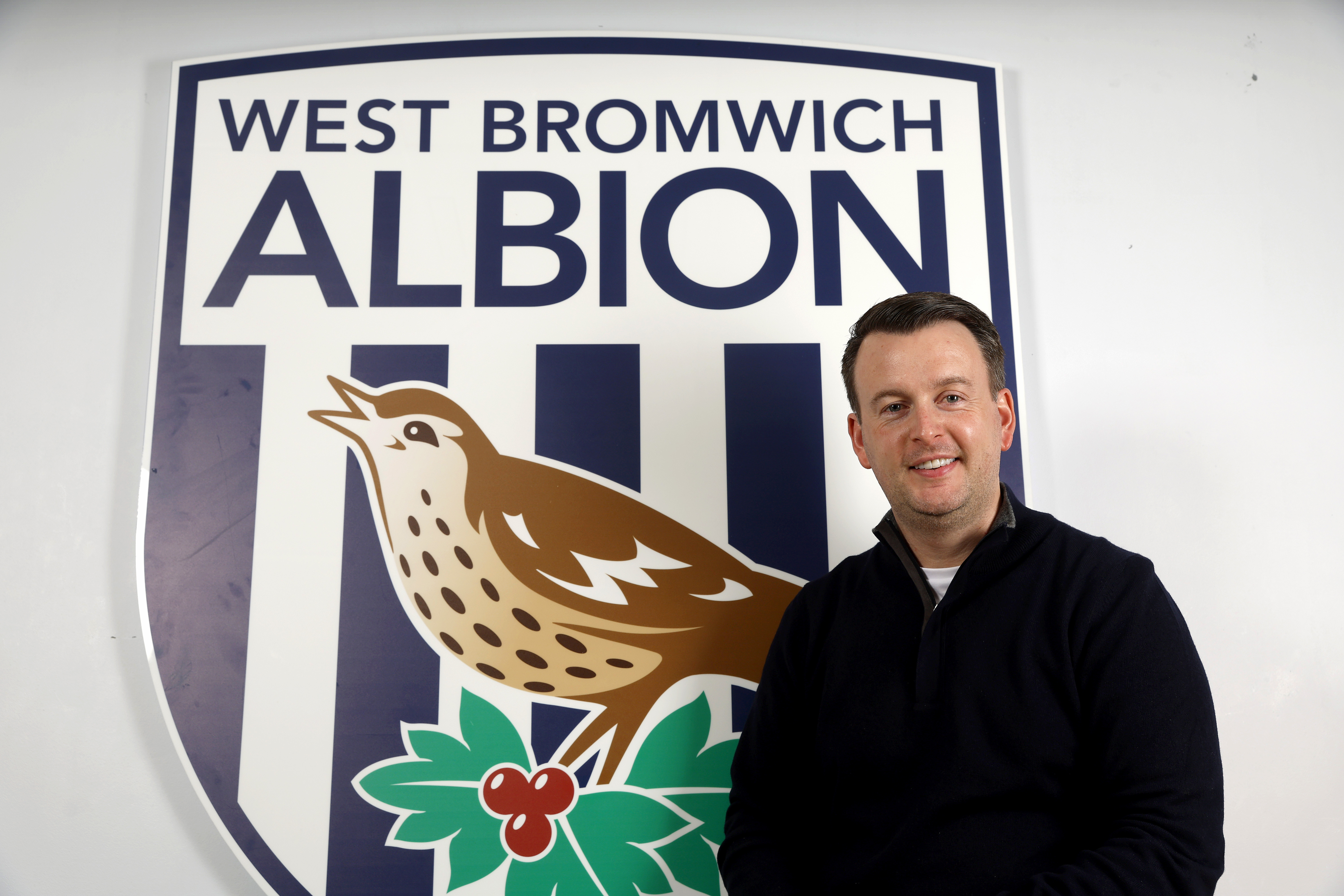 Dominic Price smiling at the camera while stood by a WBA badge 